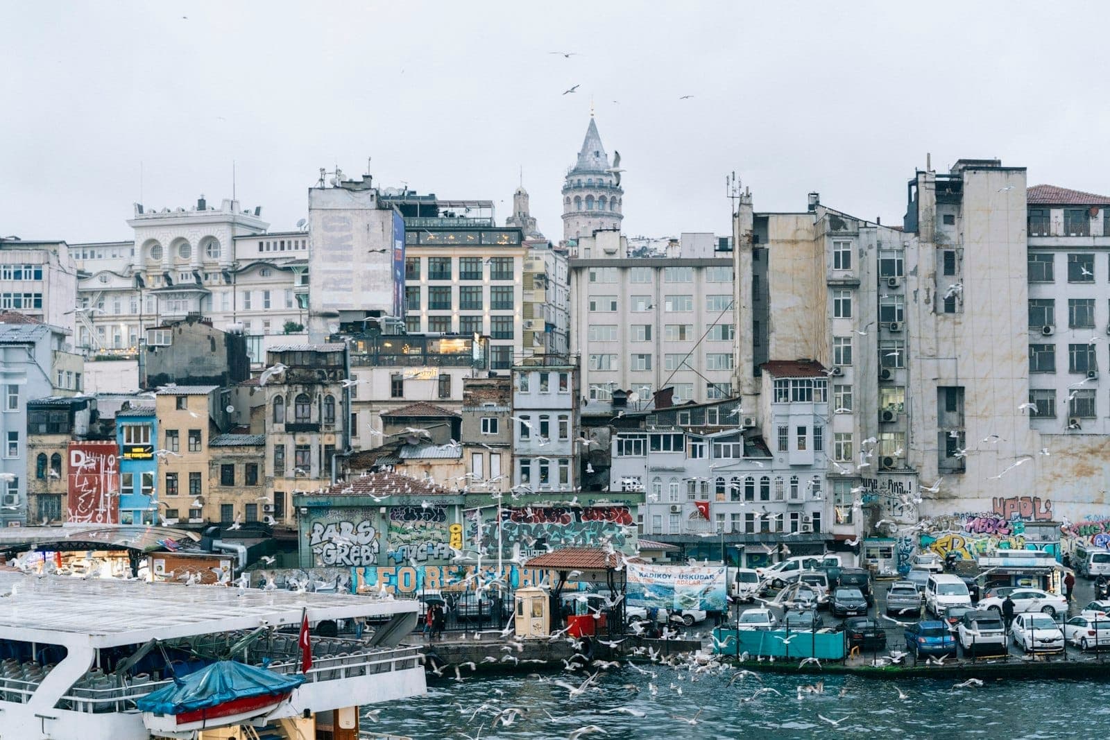 Blick auf die Uferpromenade von Karaköy mit historischen Fassaden, lebhaften Graffiti, einem angedockten Fährschiff und dem Galata-Turm, der an einem bewölkten Tag über Istanbuls Stadtlandschaft aufragt.