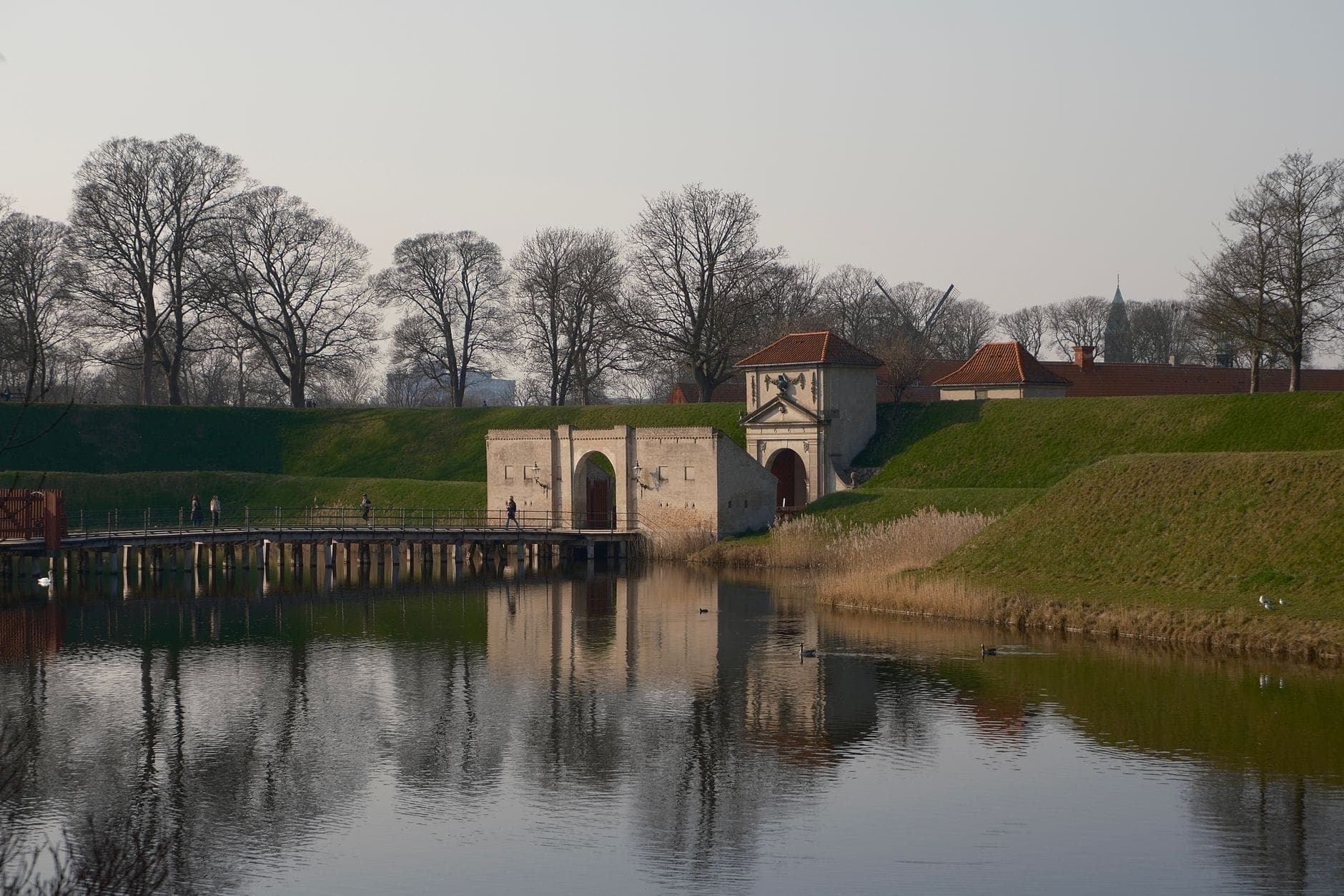 Blick auf das Eingangstor des Kastellets, den Graben und die grünen Wälle mit Bäumen, die sich an einem klaren Kopenhagener Tag im stillen Wasser spiegeln.