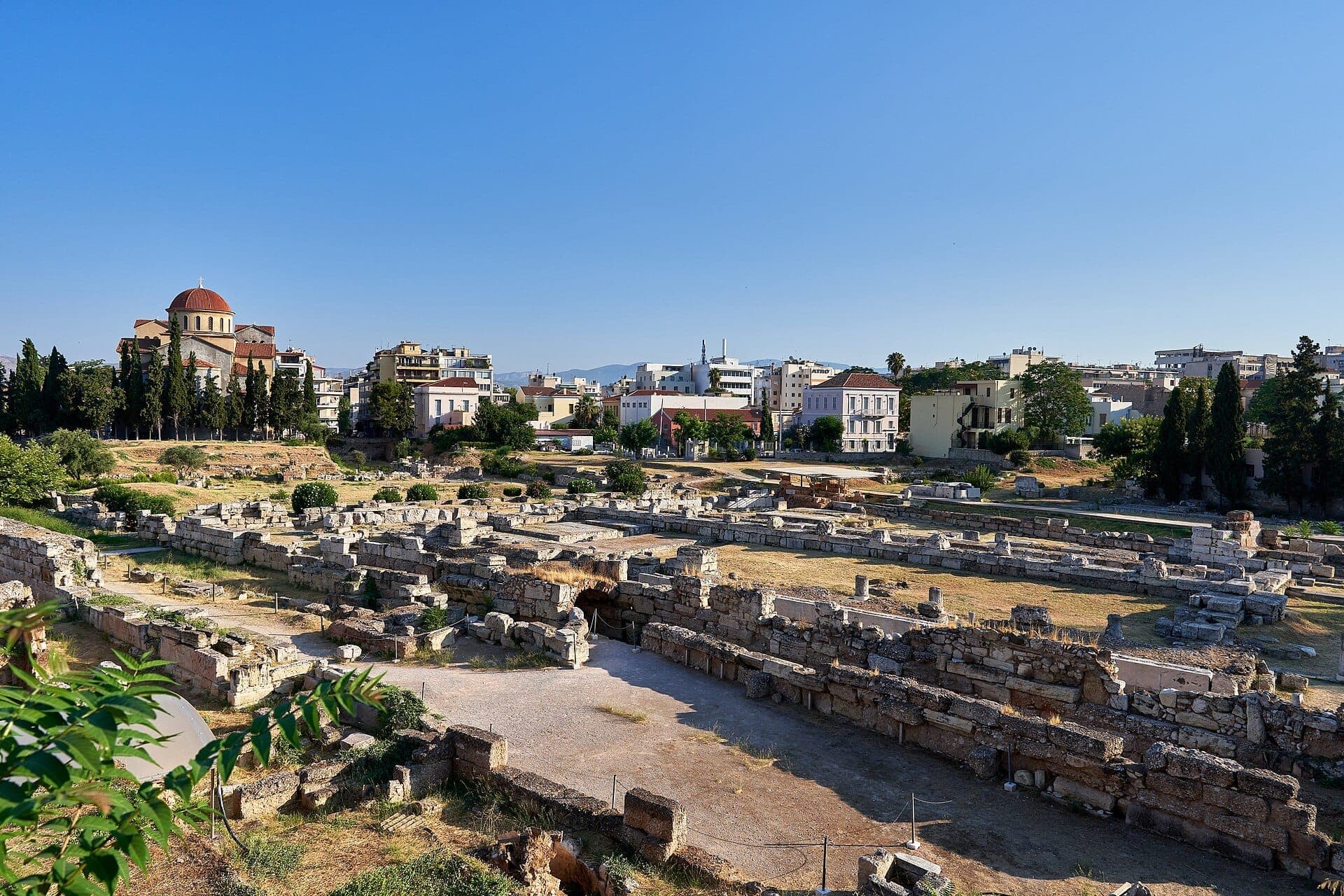 Ancient stone ruins of the Kerameikos Archaeological Site in Athens, surrounded by greenery and urban buildings under a clear blue sky.