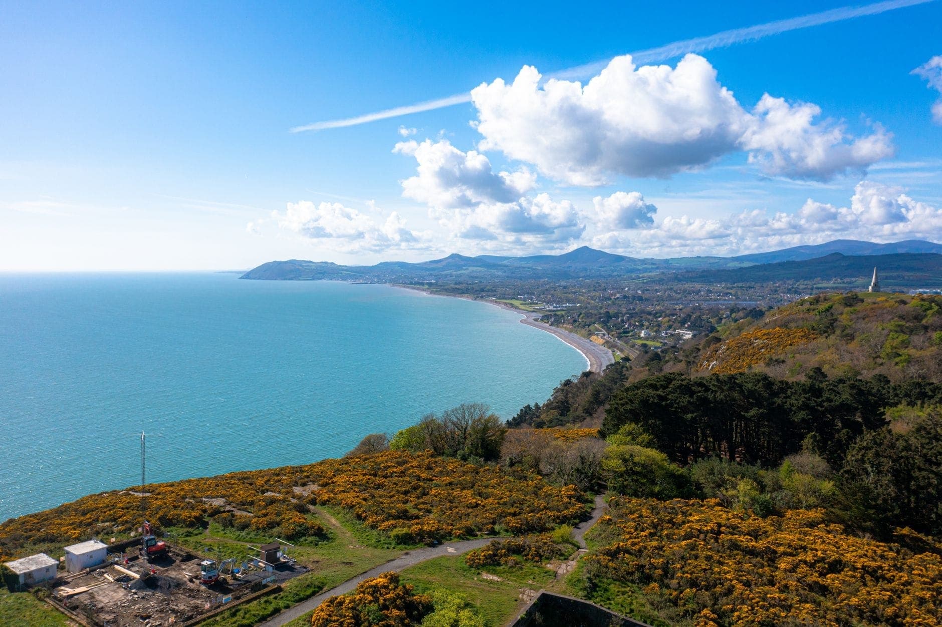 Weiter Blick vom Killiney Hill Park über die Küste, üppiges Grün, gelben Ginster, die Dublin Bay und ferne Berge unter einem strahlend blauen Himmel mit Wolken.