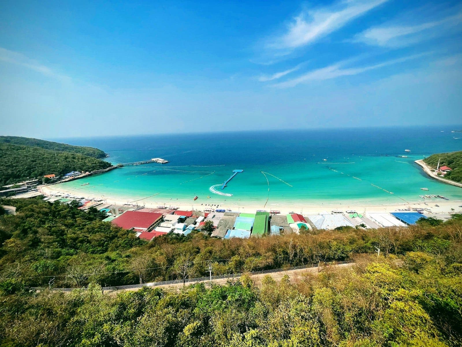 Aerial view of Koh Larn Beach showing turquoise water, white sand, lush green hills, and colorful seaside buildings under a bright blue sky.