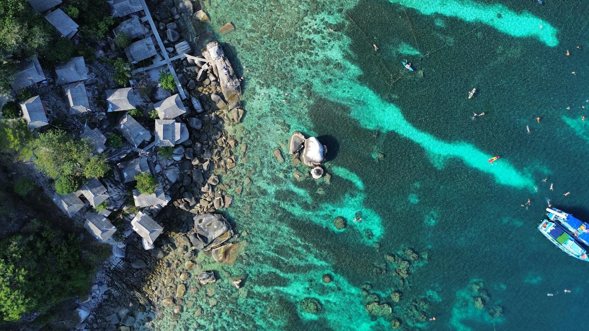 Aerial view of turquoise coral reef waters, swimmers snorkeling, boats nearby, and rustic huts along the rocky coast, showing vibrant aquatic activity.