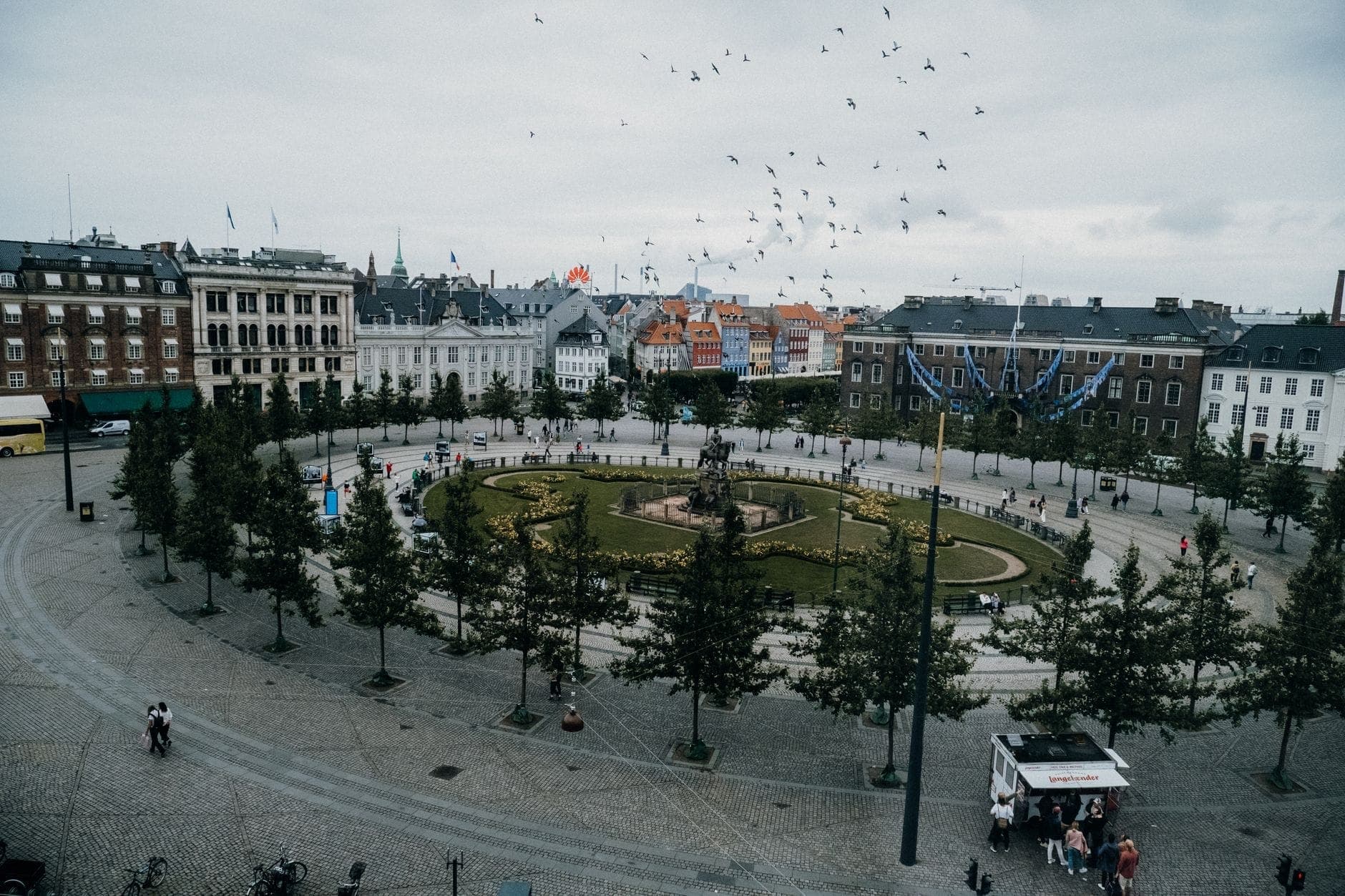 Weitwinkelperspektive auf den Kongens Nytorv in Kopenhagen mit der zentralen Statue und dem Garten, umgeben von historischen Gebäuden und Passanten.