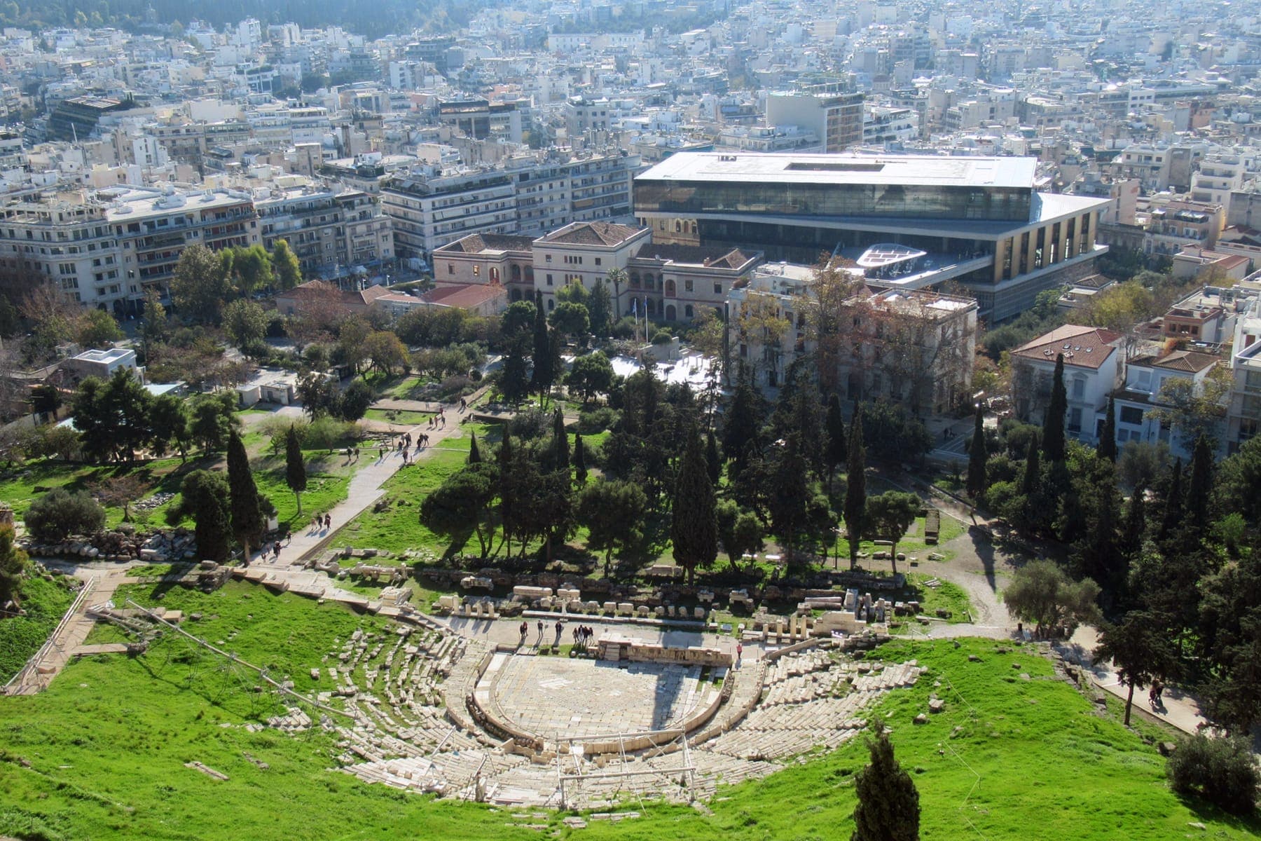 View of the ancient Theatre of Dionysus in Koukaki, with modern buildings and the Acropolis Museum in the background under clear daylight.