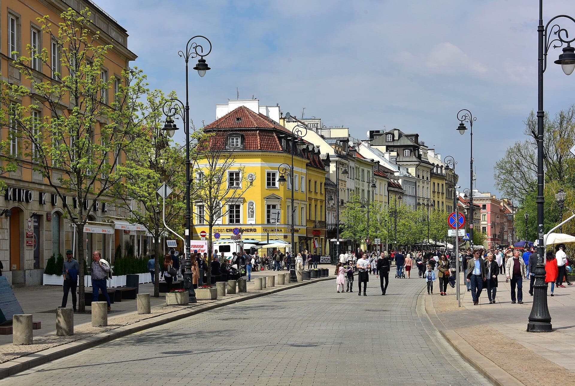 Menschen schlendern entlang der Krakowskie Przedmieście in Warschau, gesäumt von bunten historischen Gebäuden, Straßenlaternen, grünen Bäumen und belebten Straßencafés.