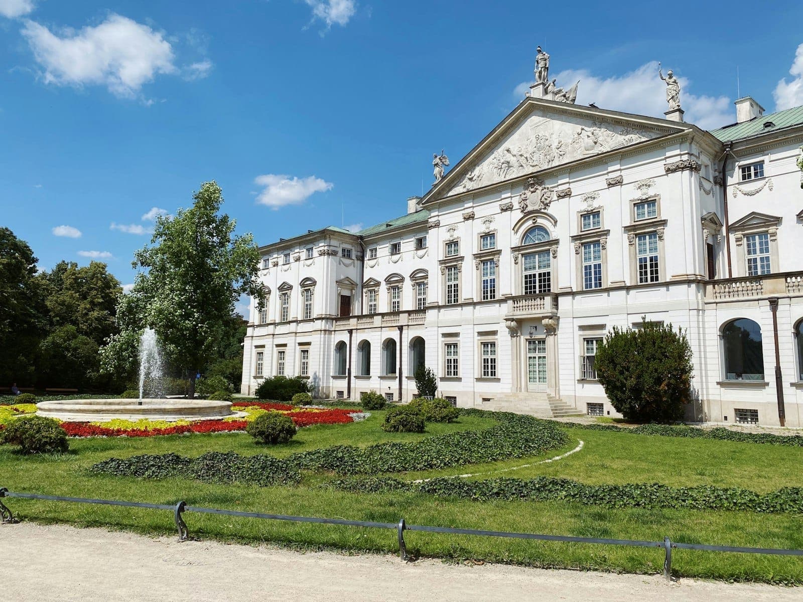 Krasiński-Palast in Warschau mit seiner weißen Barockfassade, üppigen grünen Gärten, bunten Blumenbeeten und einem zentralen Brunnen unter einem strahlend blauen Himmel.