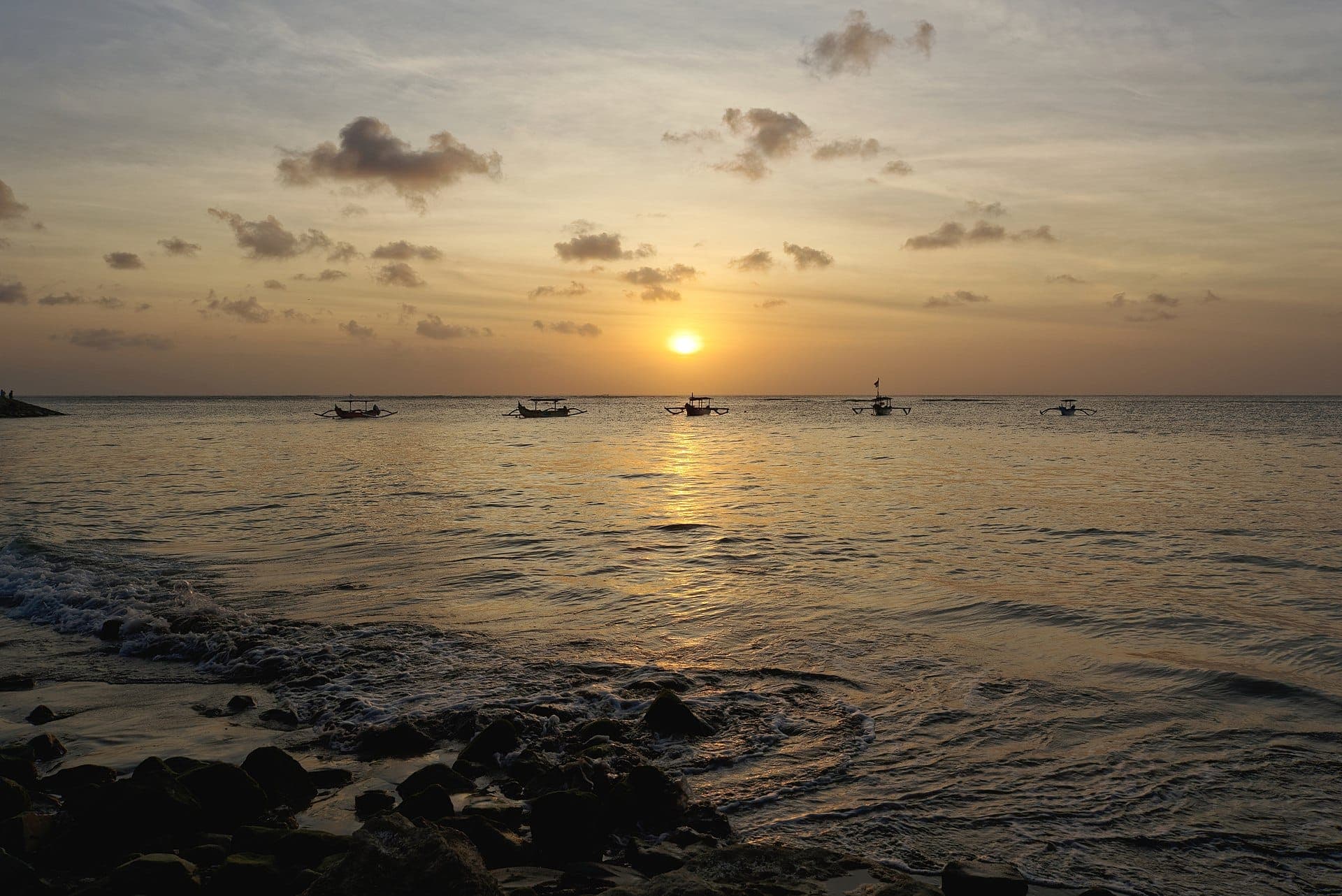 Toàn cảnh hoàng hôn nhìn từ Kuta Beach, Bali, với những con sóng nhẹ nhàng và những chiếc thuyền xa xa ở đường chân trời.