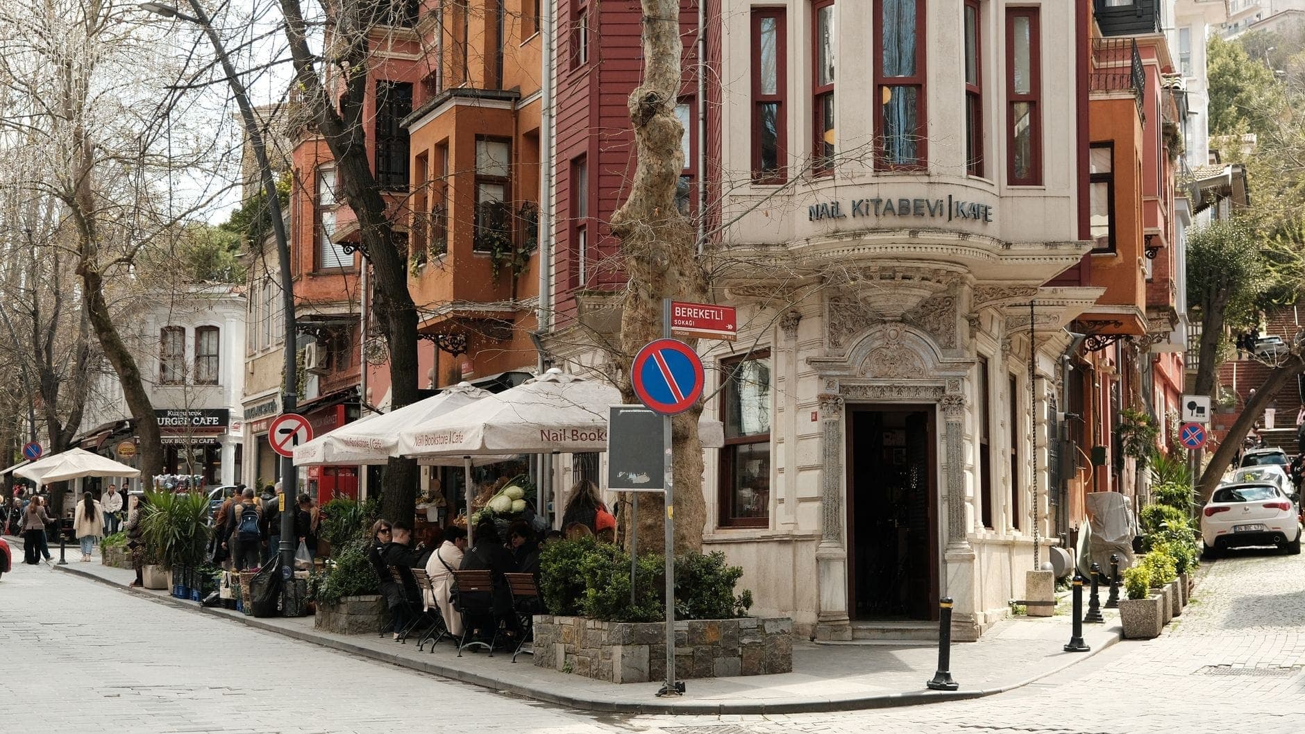 Straßenansicht von Kuzguncuk mit pastellfarbenen Holzhäusern, Café-Bestuhlung im Freien und entspannten Einheimischen in einer lebhaften, charmanten Atmosphäre.