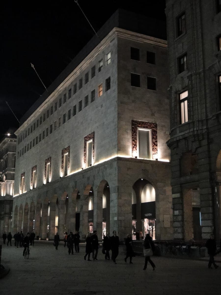 La Rinascente Milan department store illuminated at night, with arched stone façade and evening shoppers walking along Piazza del Duomo.