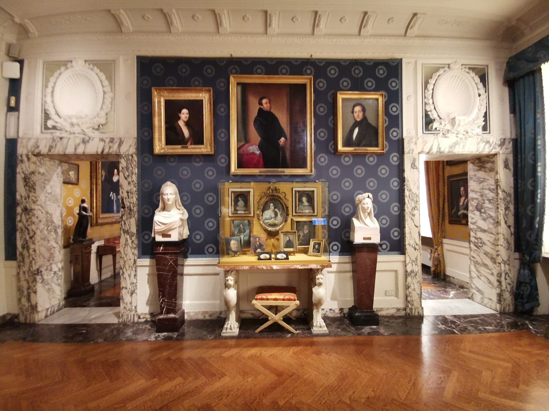 Gallery inside Museo Teatrale alla Scala featuring marble pedestals, classical busts, ornate portraits, and elegant blue patterned walls with natural lighting.