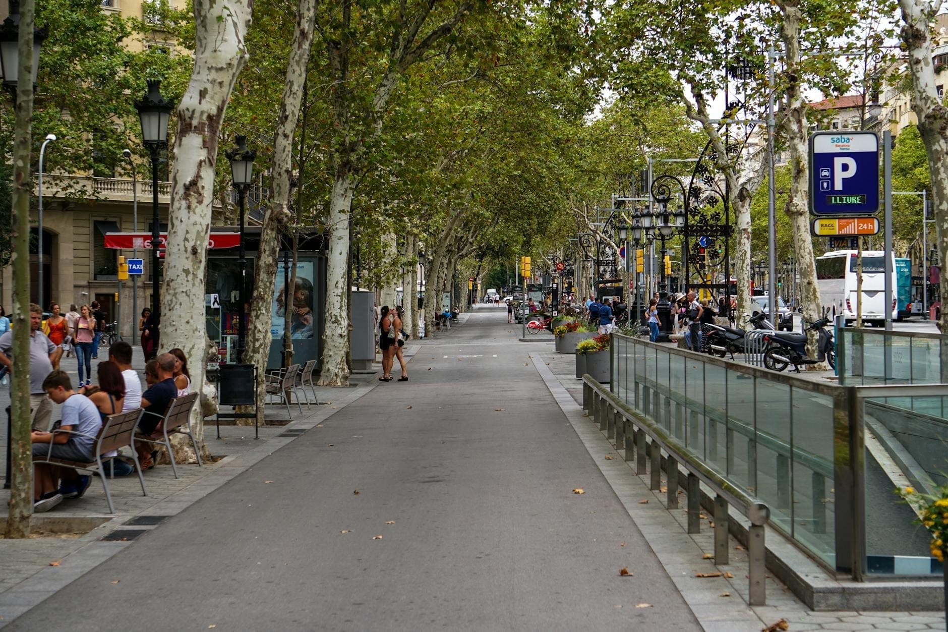 Ang malawak na abenida ng Las Ramblas sa Barcelona, may mga puno sa magkabilang gilid, mga tao sa labas ng mga kainan, at mga manlalakad sa gitna ng daan.