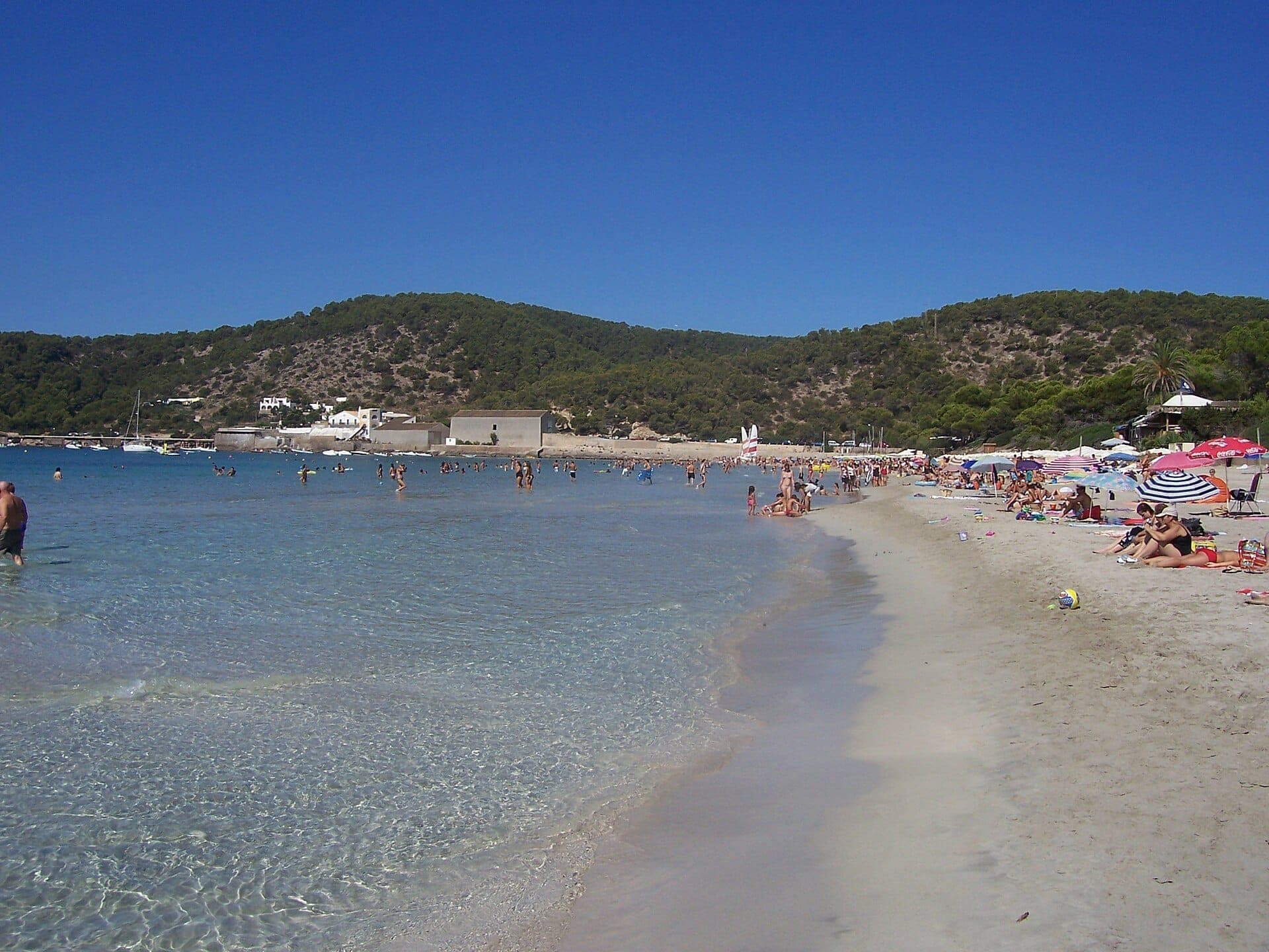 Crowds relax under colorful umbrellas along the clear shoreline of Platja de Ses Salines, with pine-covered hills and white buildings in the background.