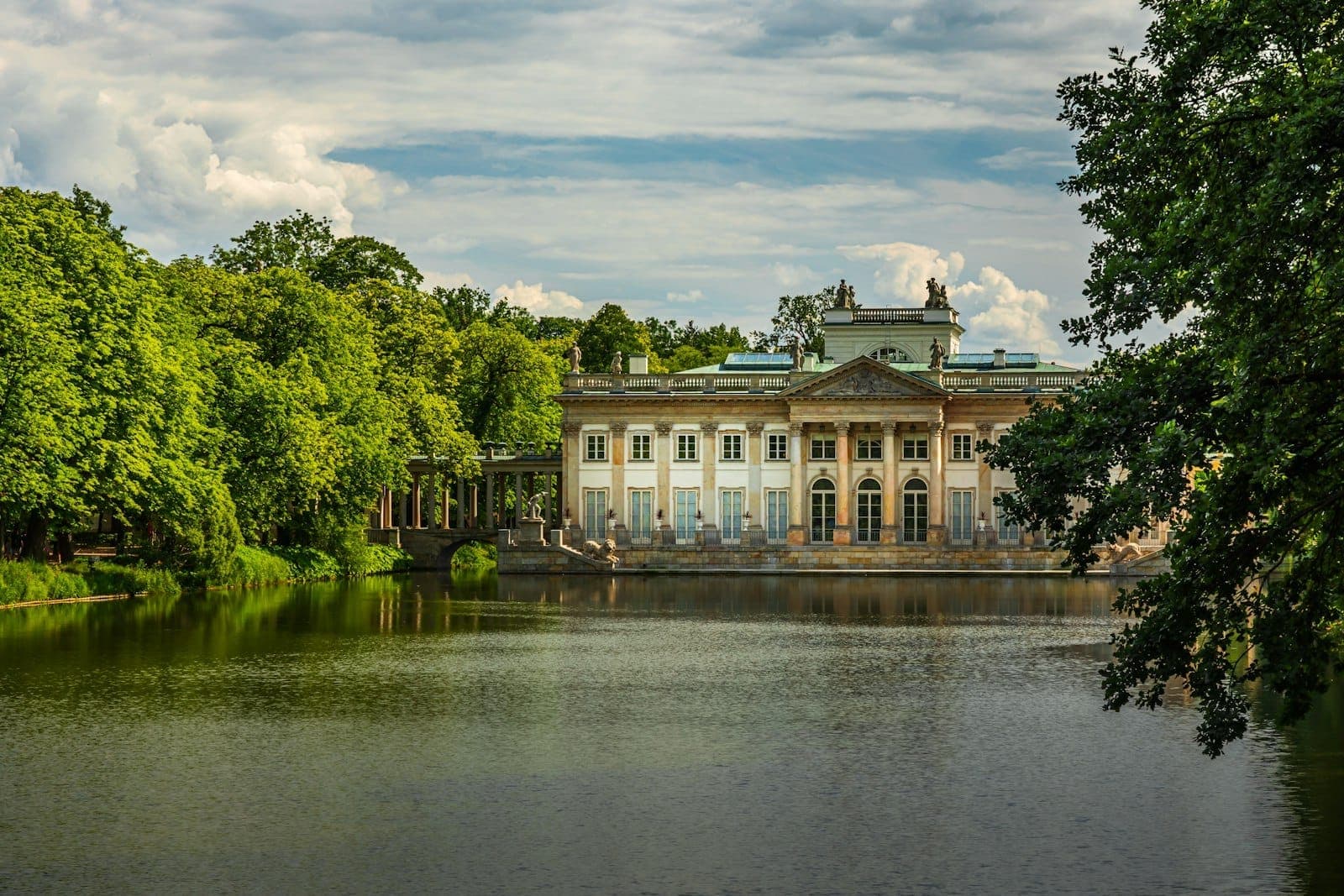 Ein klassizistischer Palast spiegelt sich in einem stillen See, umgeben von üppigen grünen Bäumen an einem hellen Tag im Łazienki-Park, Warschau.