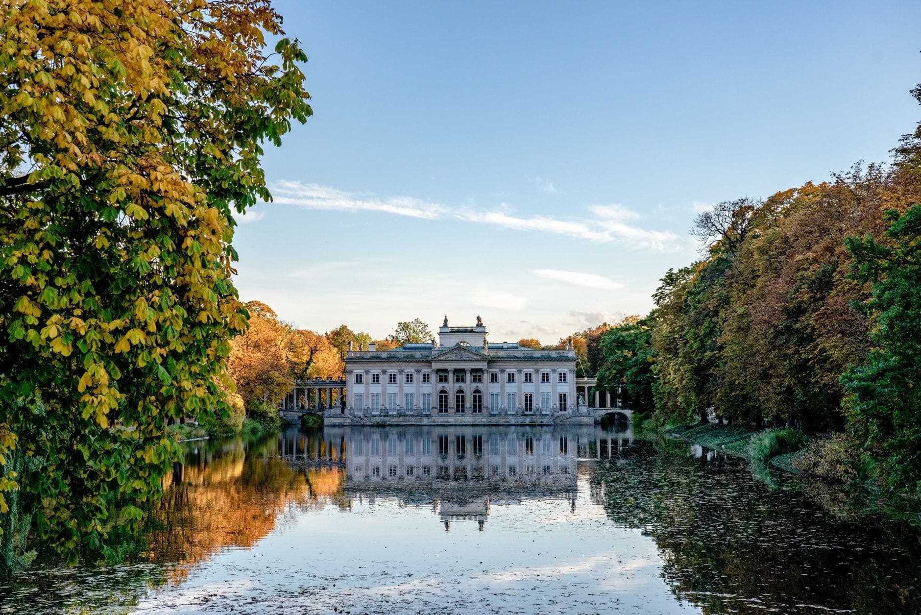 Weitläufiger Blick auf den Palast auf der Insel im Łazienki Park, der sich im See spiegelt, eingerahmt von üppigen Bäumen in frühen Herbstfarben unter einem strahlend blauen Himmel.