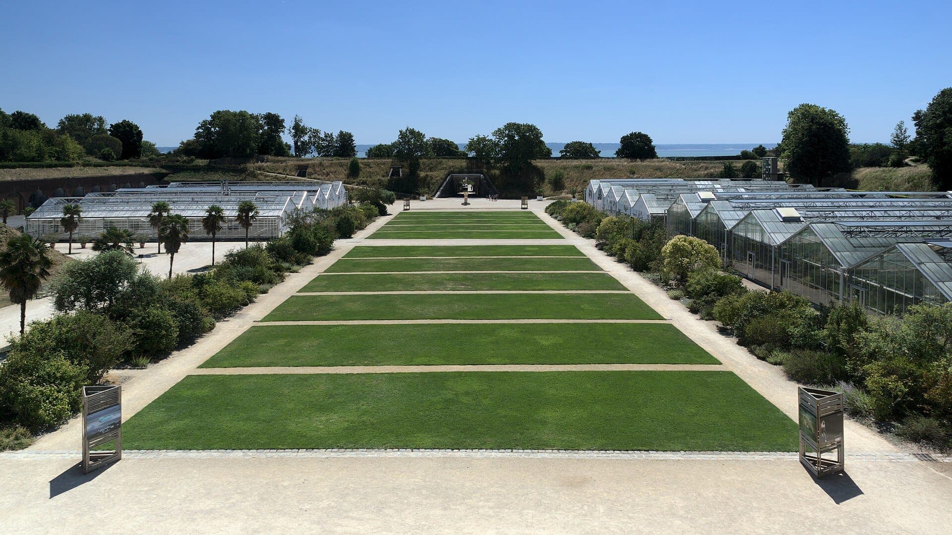 Bãi cỏ trung tâm tại Les Jardins Suspendus ở Le Havre, hai bên là nhà kính và các luống cây xanh tươi tốt dưới bầu trời xanh trong.