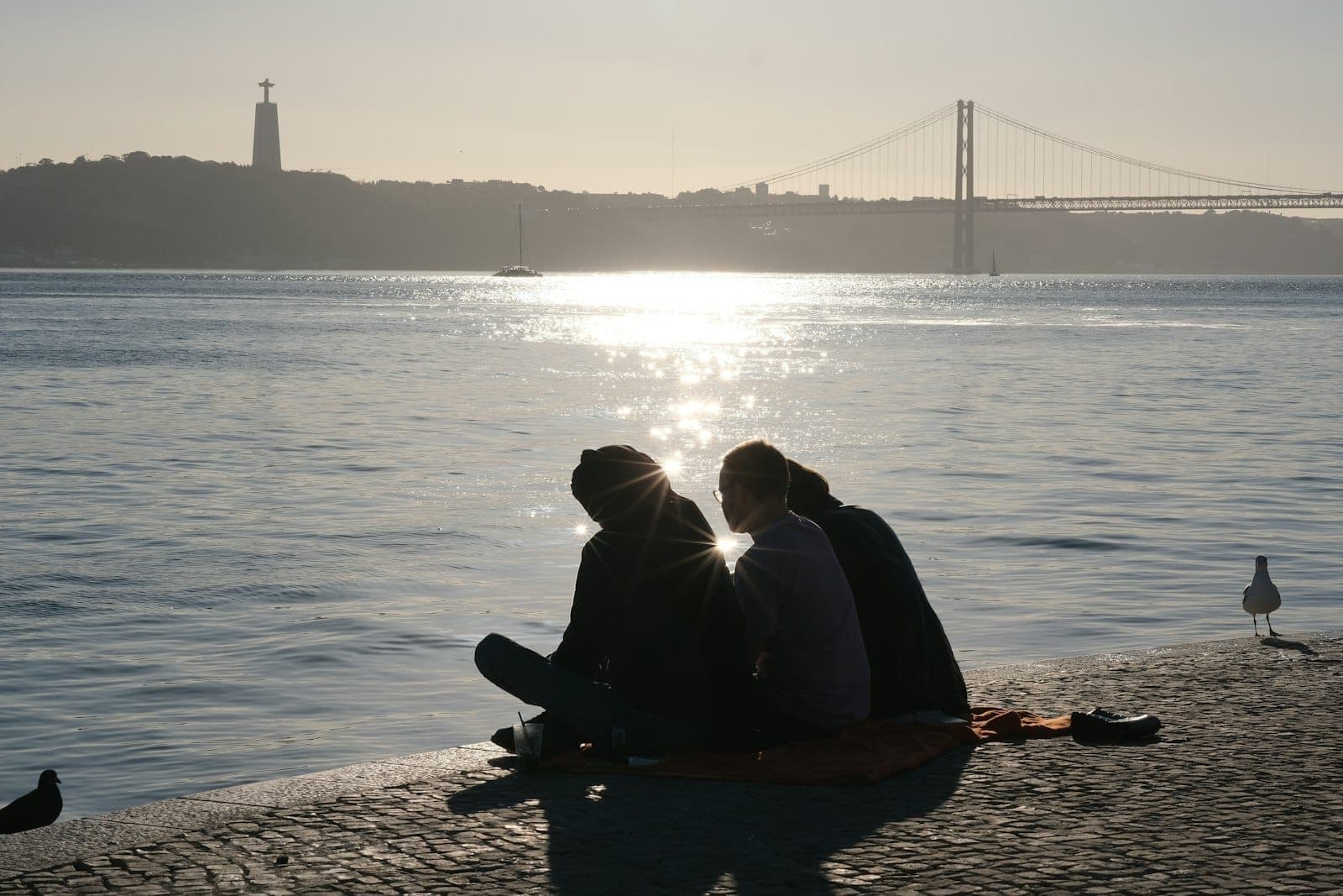 Cặp đôi ngồi sát nhau bên bờ sông lúc hoàng hôn ở Lisbon, với cầu 25 de Abril và tượng Cristo Rei phía sau.