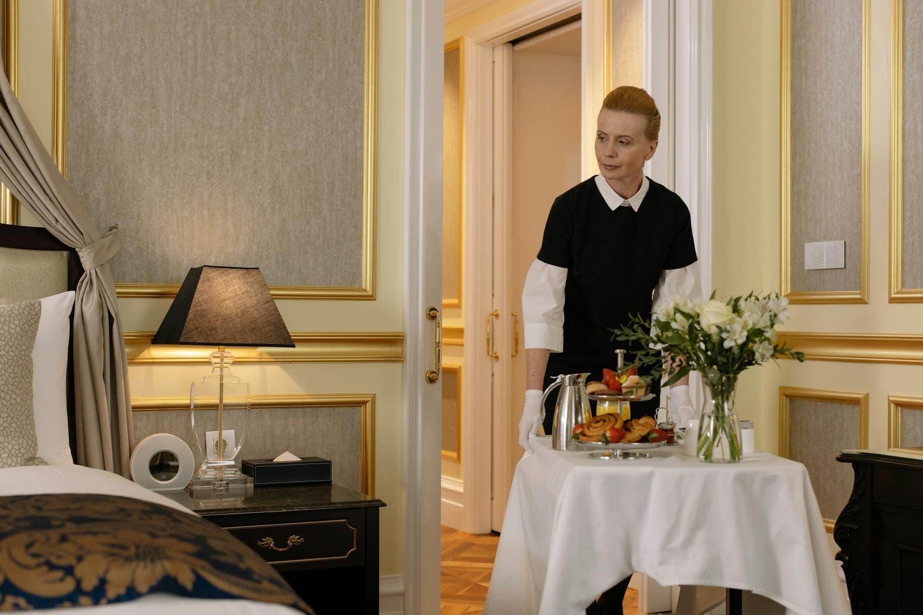 Phòng suite khách sạn sang trọng với điểm nhấn vàng thanh lịch, nhân viên ăn mặc chỉnh tề đang phục vụ bữa sáng trên khay, không gian ấm cúng và đẳng cấp.