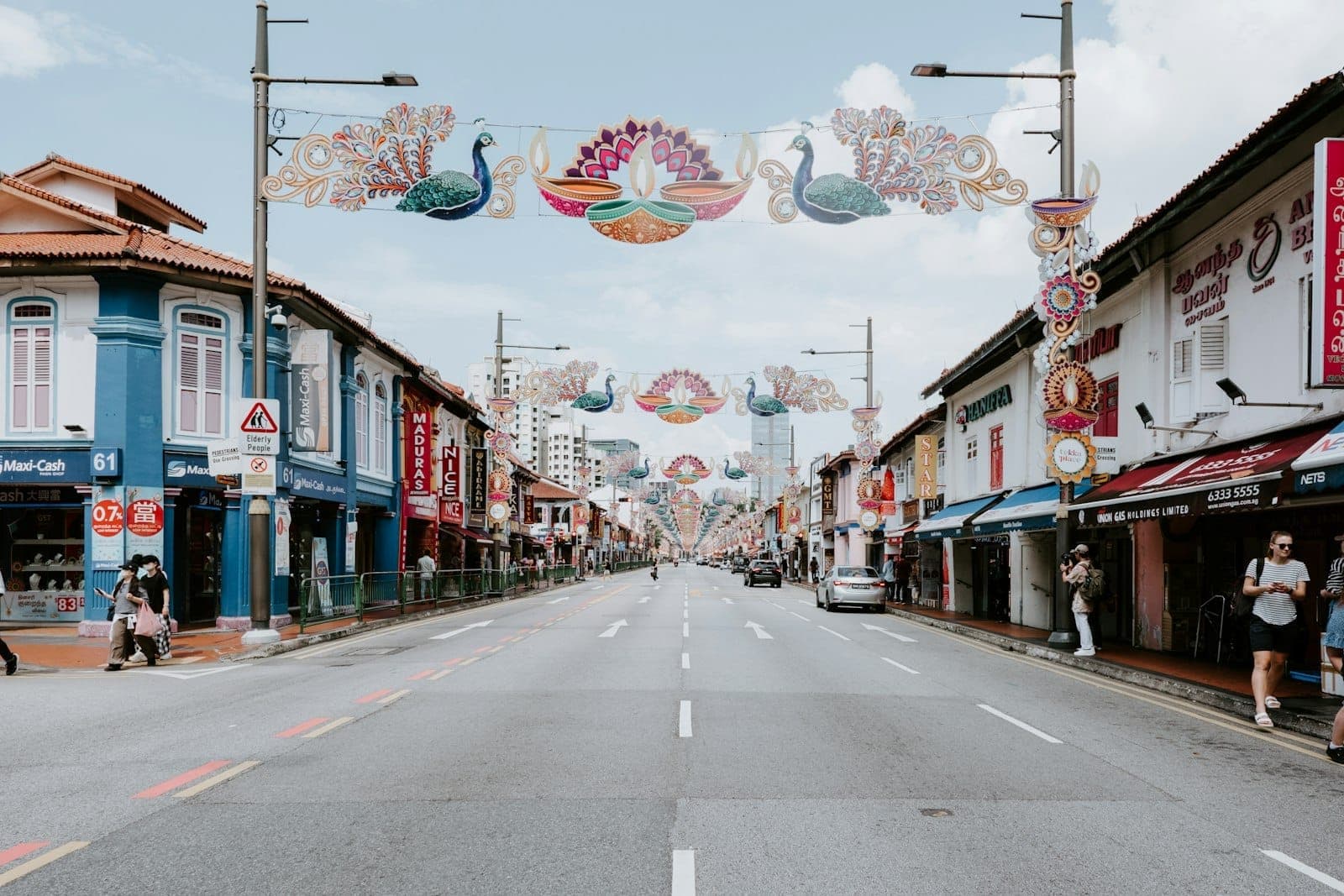 Wide view of Serangoon Road in Little India, Singapore, with colorful heritage shophouses and ornate peacock-themed festive street decorations under a bright sky.