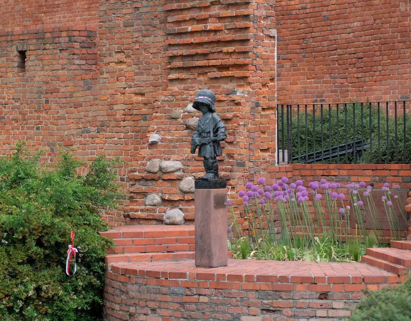 Die Bronze-Statue des Kleinen Aufständischen zeigt einen kindlichen Soldaten, der ernst vor den historischen roten Backsteinmauern der Warschauer Altstadt steht, umgeben von grünen Sträuchern und lila Blumen.