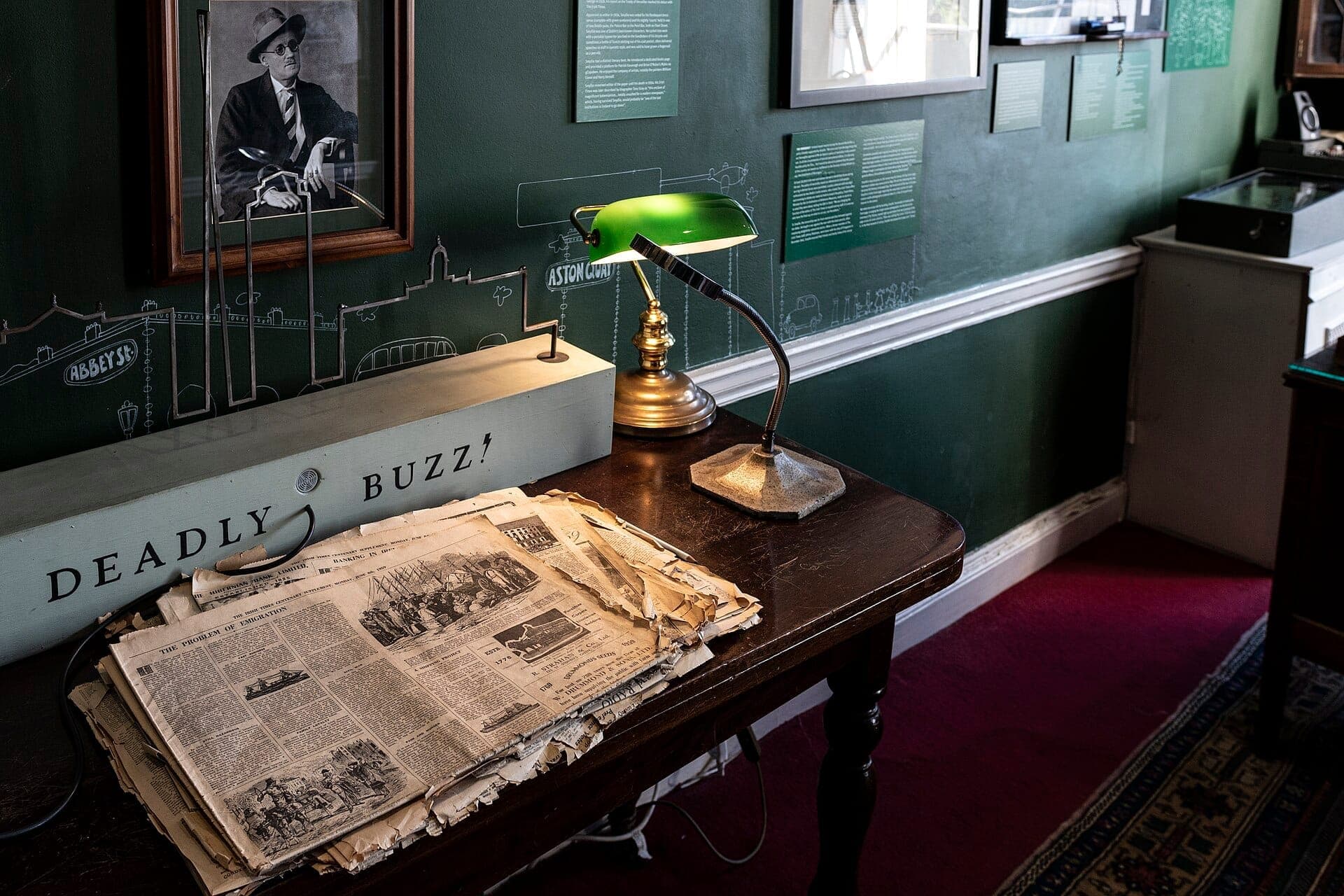 Ekspozycja w Little Museum of Dublin przedstawiająca stare gazety, zieloną lampę bankierską i historyczne artefakty na ciemnym drewnianym biurku.