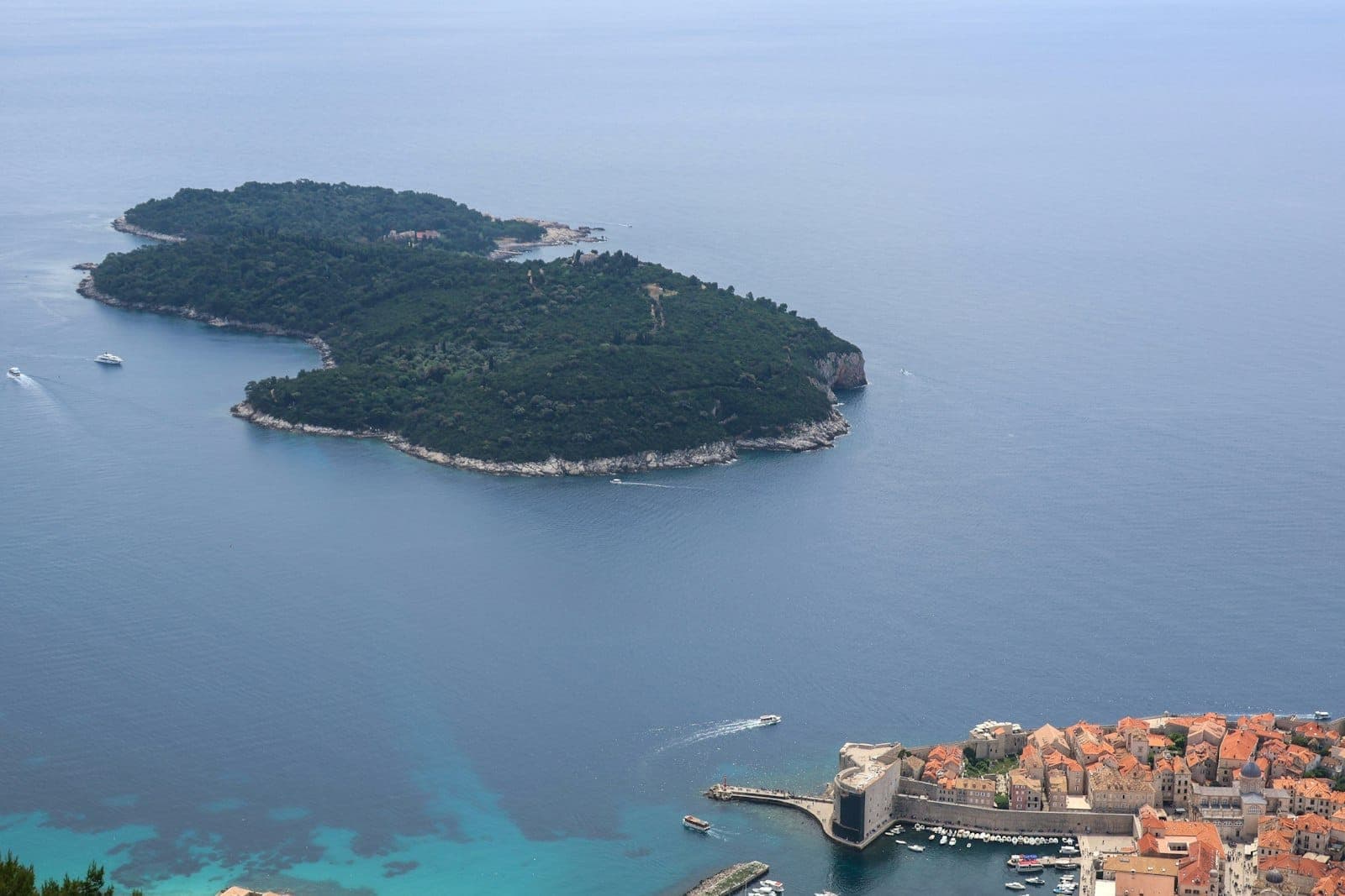 Aerial na tingin ng Isla ng Lokrum na napalibutan ng asul na dagat, na may Lumang Lungsod at daungan ng Dubrovnik sa harap sa isang maliwanag na araw.