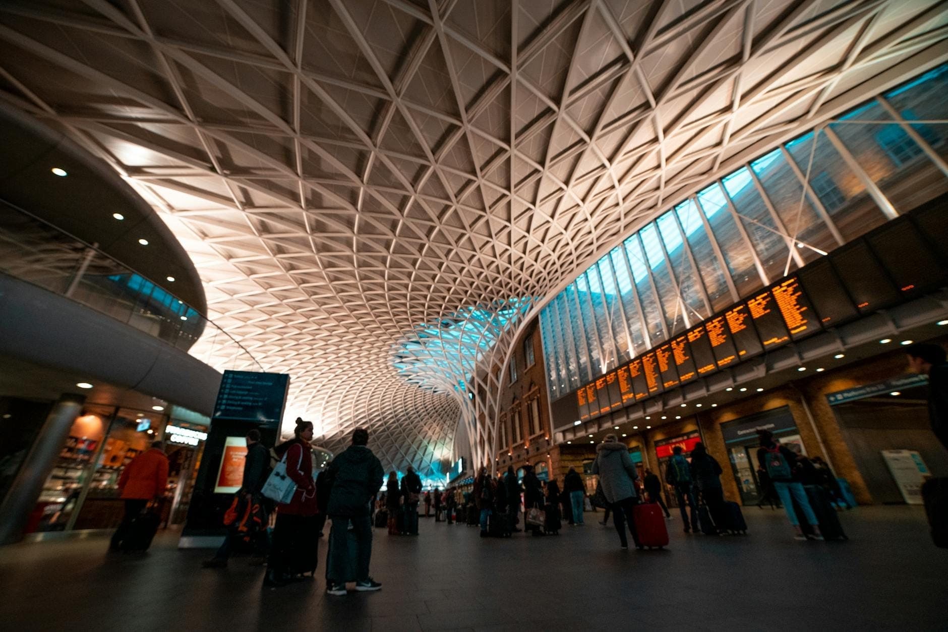 Inside London King's Cross station, travelers stand under an intricate modern roof with a large departures board visible overhead.