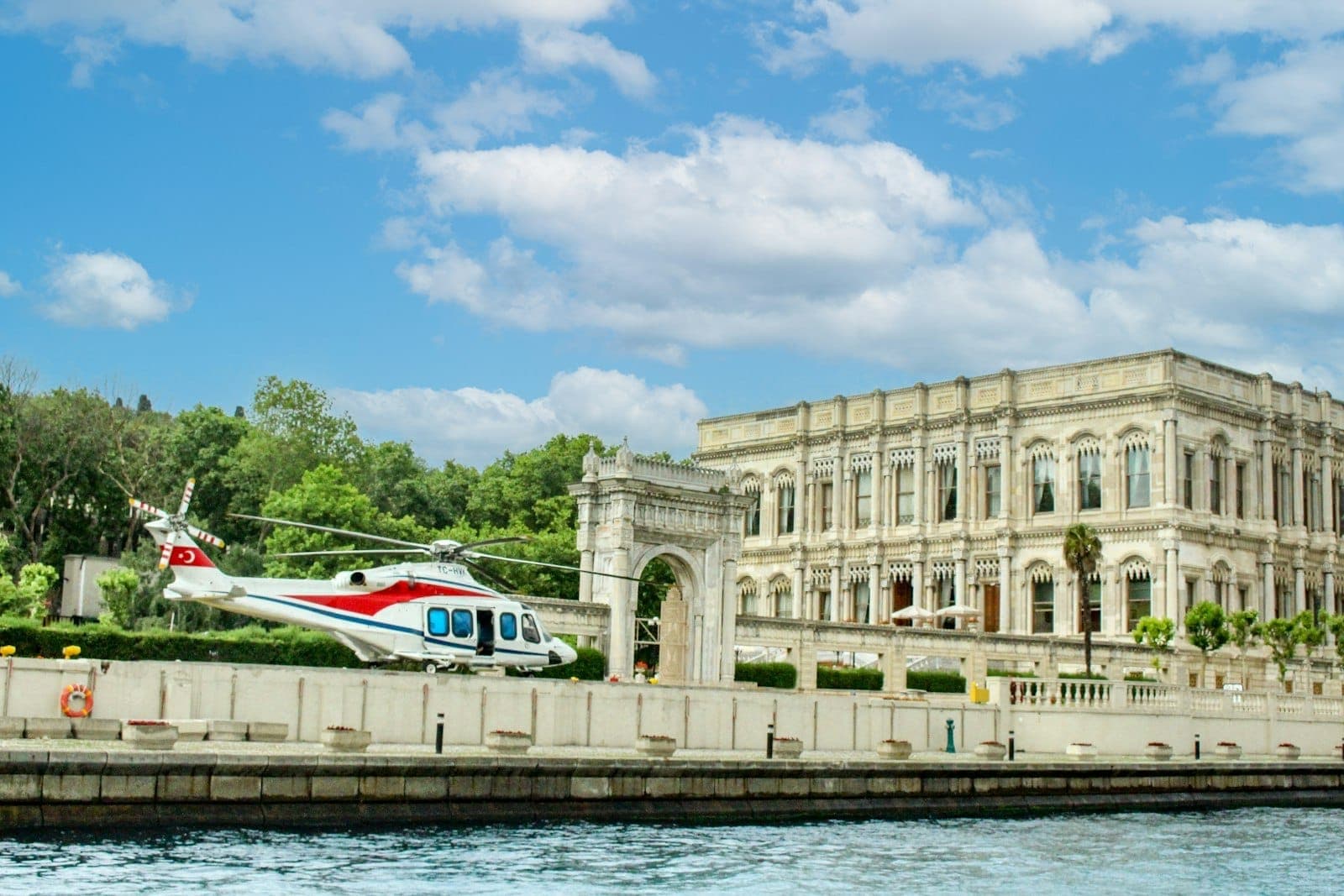 Eleganter neoklassizistischer Palast direkt am Bosporus mit üppigen Gärten und einem privaten Hubschrauber auf dem Landeplatz, unter strahlend blauem Himmel mit weißen Wolken.