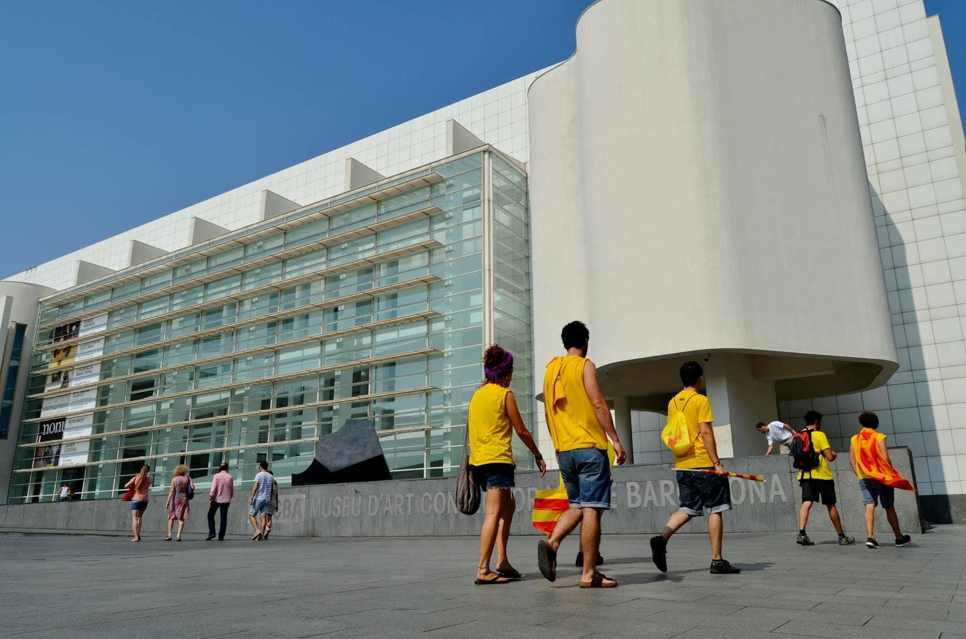 Bên ngoài MACBA ở Barcelona, nổi bật với mặt tiền trắng hiện đại, tường kính và các nhóm người đang đi bộ trong quảng trường ngập nắng.