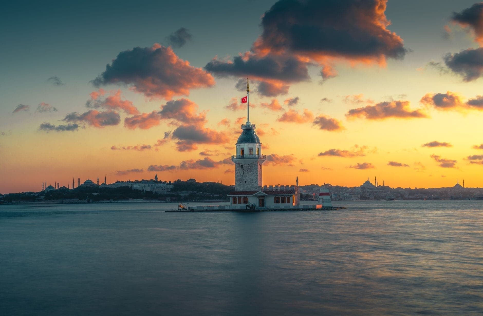Der Kız Kulesi steht allein auf seinem Eiland im Bosporus bei Sonnenuntergang – dramatischer Himmel, Istanbuls Skyline in der Ferne.