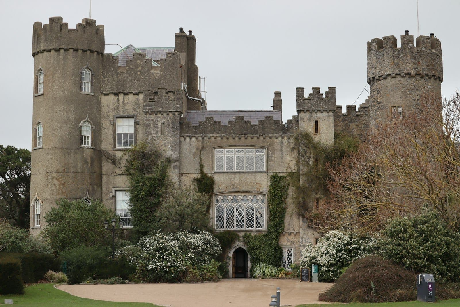Front view of Malahide Castle with its iconic round towers, medieval stone facade, arched entrance, and surrounding lush gardens in spring blooms.