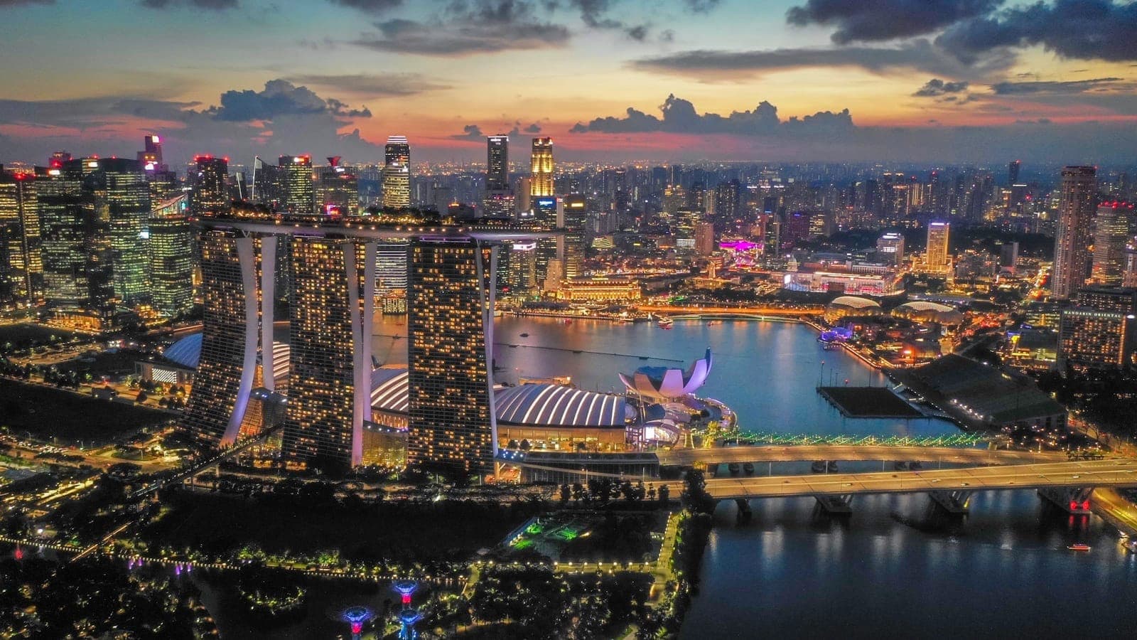 Aerial evening view of Marina Bay with Marina Bay Sands, city skyline, waterfront, and vibrant city lights under a dramatic sunset sky.