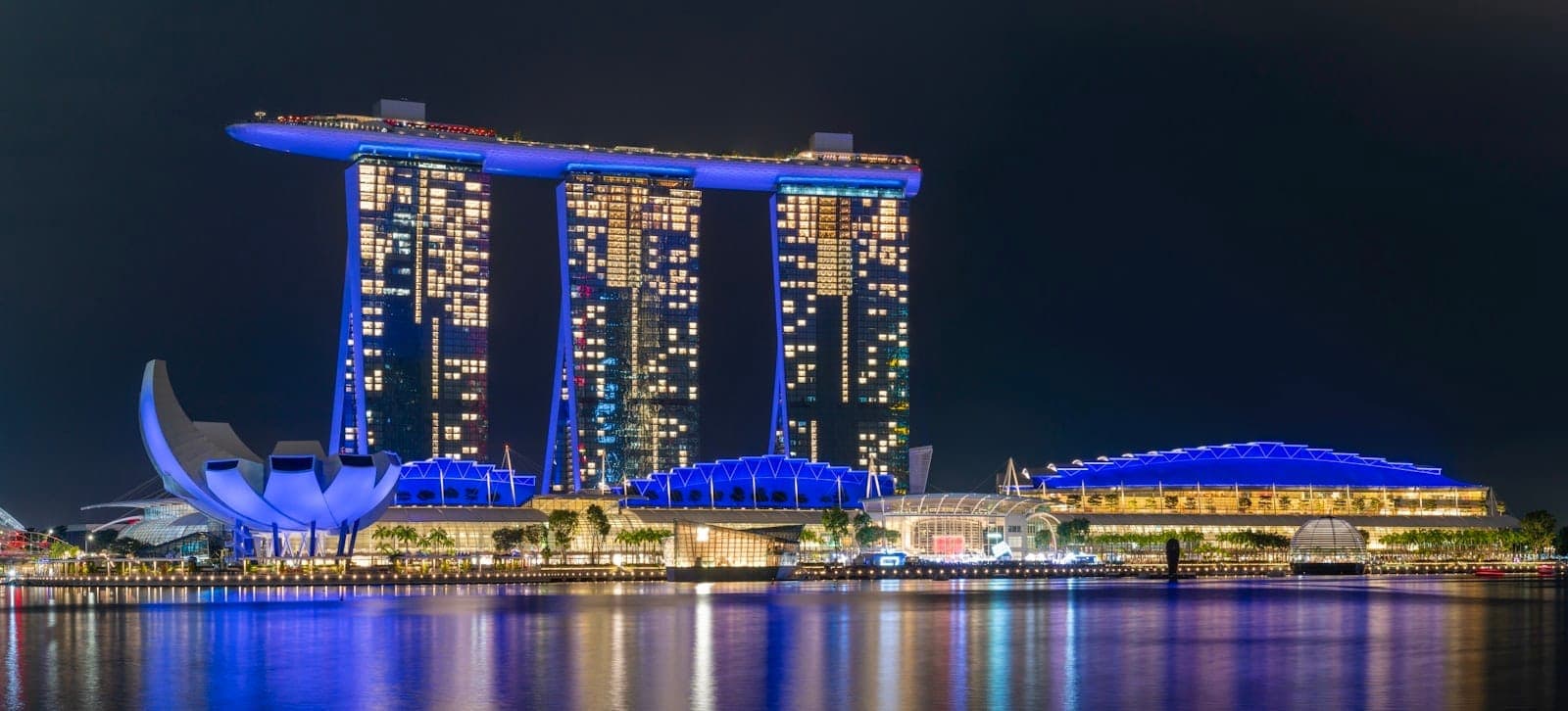 Nighttime panoramic view of Marina Bay with Marina Bay Sands, ArtScience Museum, and illuminated waterfront promenade reflected in calm bay waters.