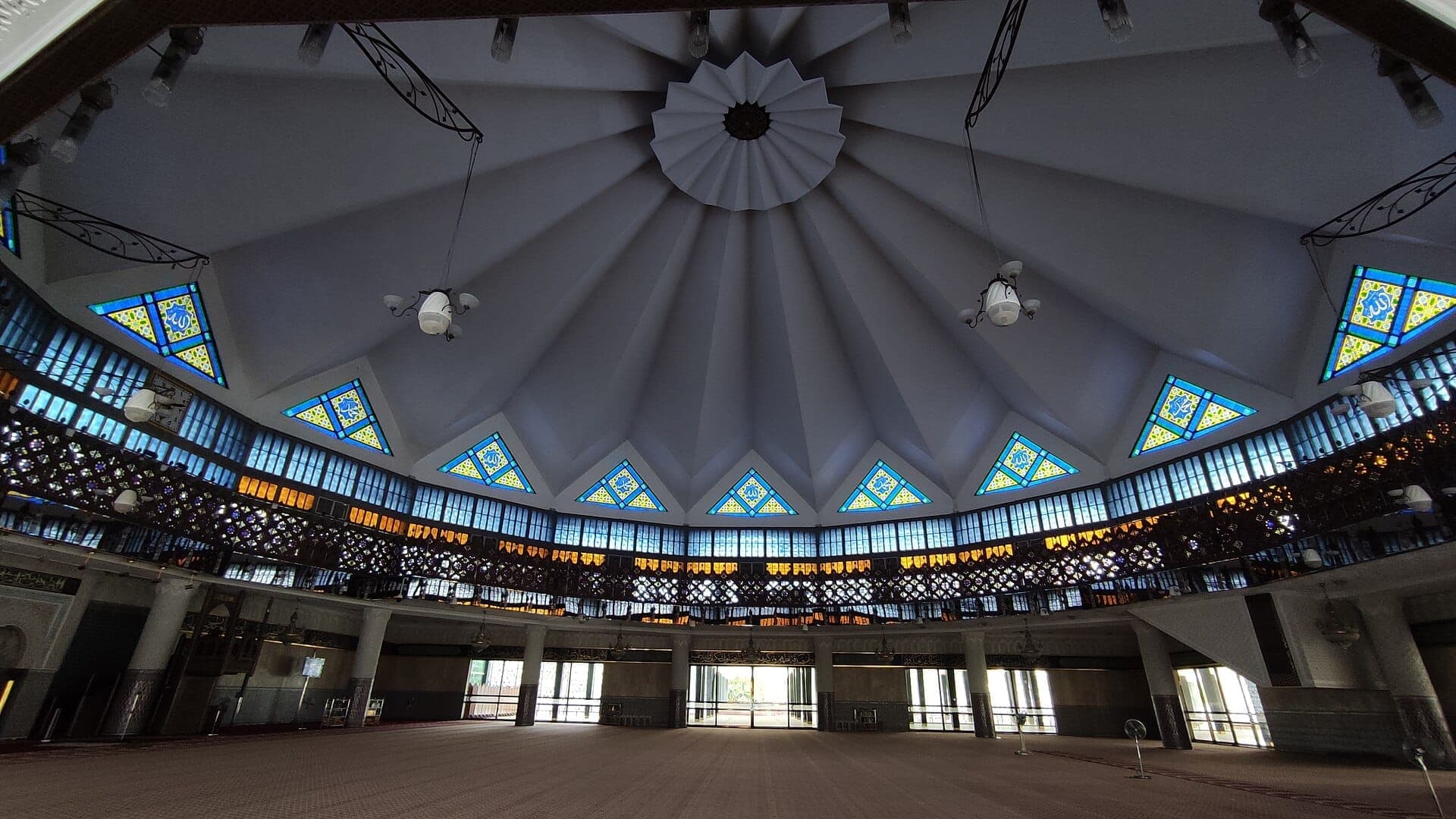 Cảnh bên trong Masjid Negara với mái vòm theo phong cách hiện đại nổi bật, cửa sổ kính màu hình học và phòng cầu nguyện mở được chiếu sáng bởi ánh sáng tự nhiên.