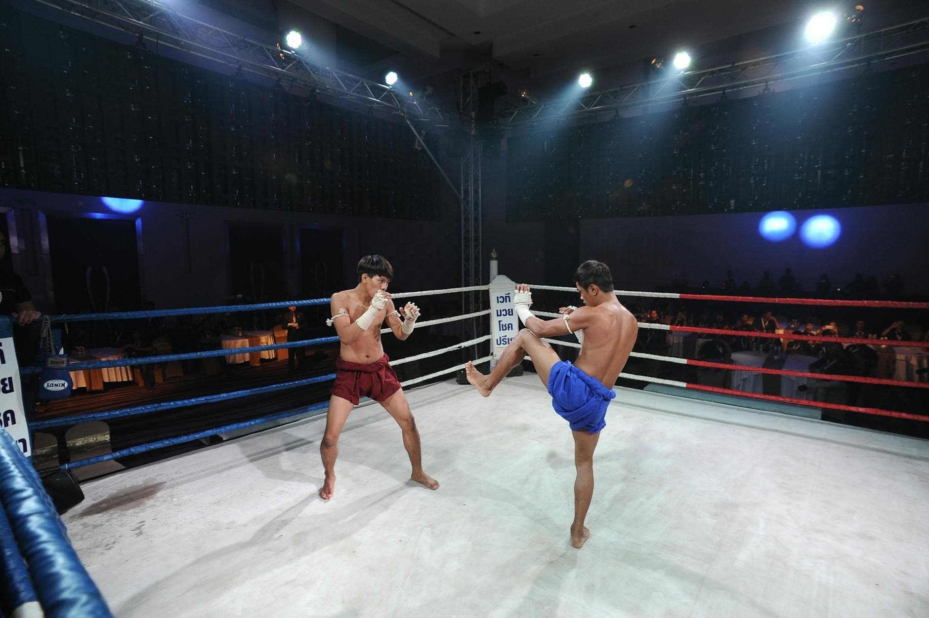 Two Muay Thai fighters in red and blue shorts square off in a brightly lit ring, with an excited audience watching in the background.
