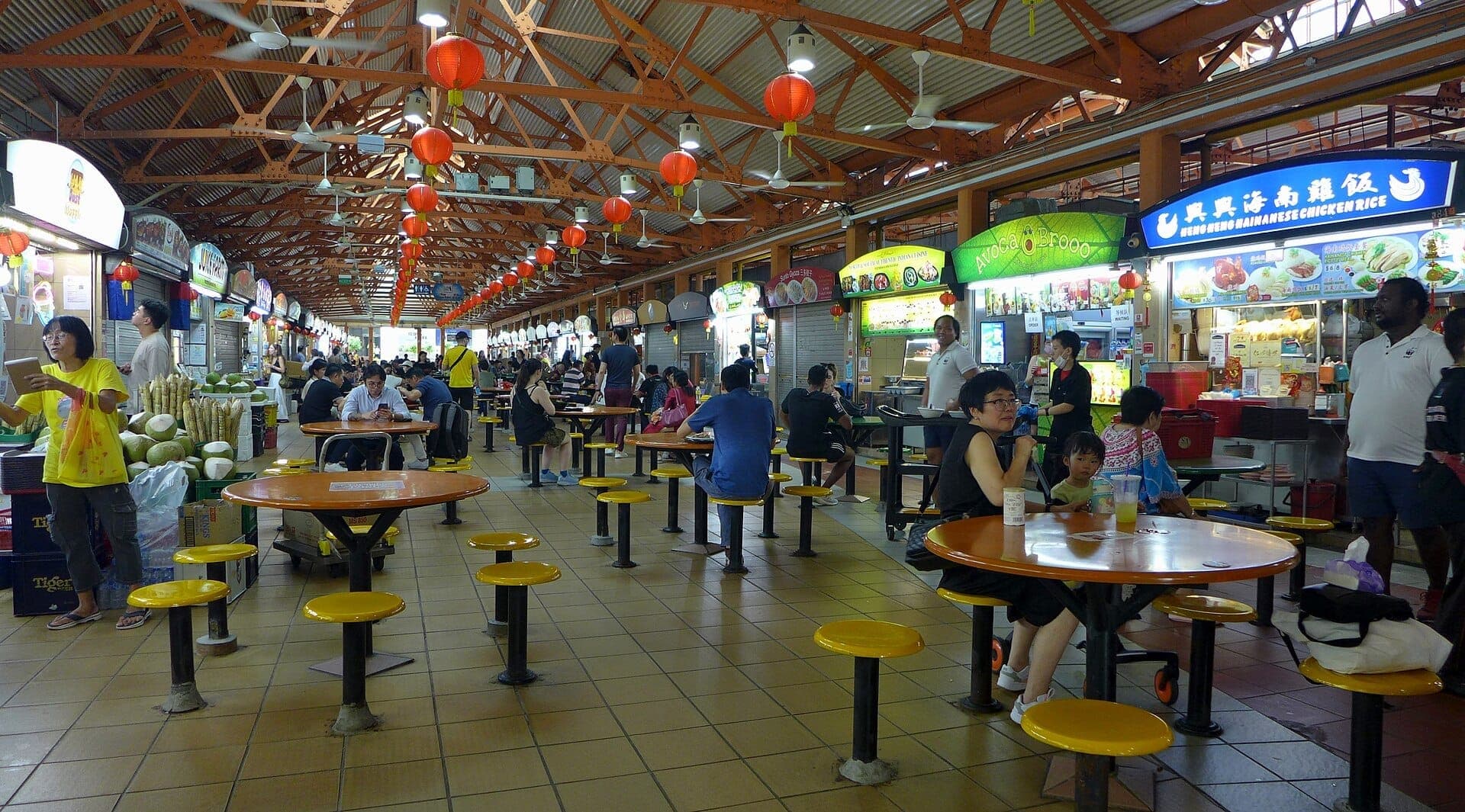 Maxwell Food Centre interior with rows of food stalls, orange tables, yellow stools, and diners enjoying meals under red lanterns and open ceiling beams.