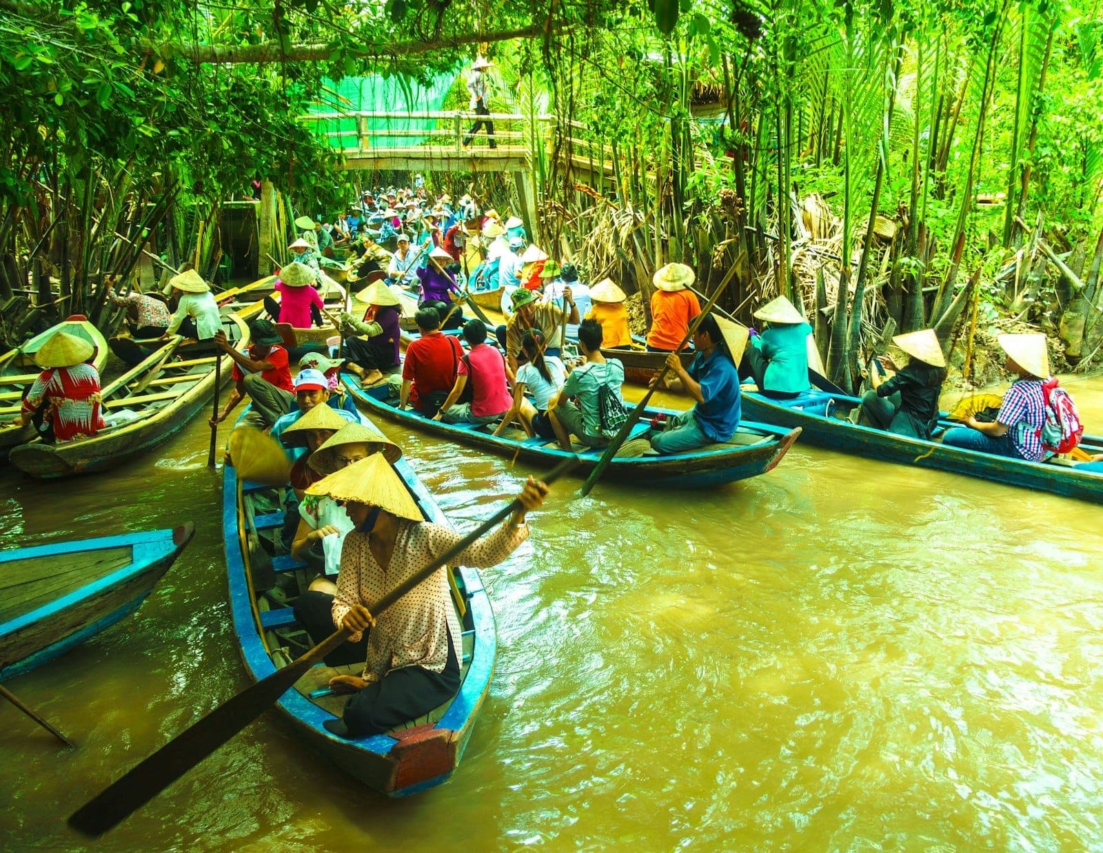 Du khách đội nón lá chèo thuyền nhỏ trên con kênh bùn đỏ, hai bên là cây xanh nhiệt đới rậm rạp ở Đồng bằng Sông Cửu Long.