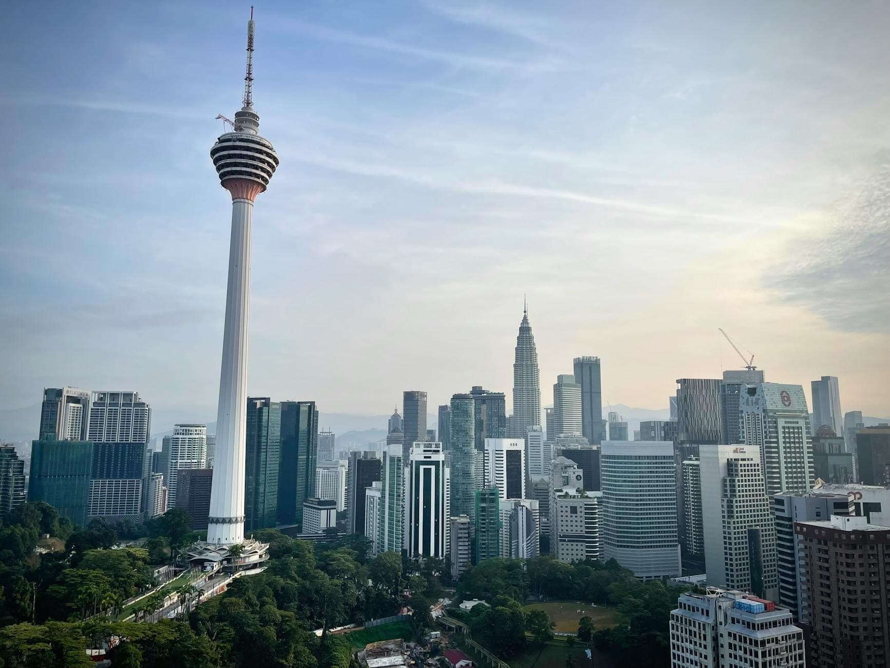 Tháp Menara KL Tower sừng sững vươn cao trên đường chân trời Kuala Lumpur, bao quanh là những tòa nhà chọc trời hiện đại và cây xanh tươi tốt dưới bầu trời có mây rải rác.