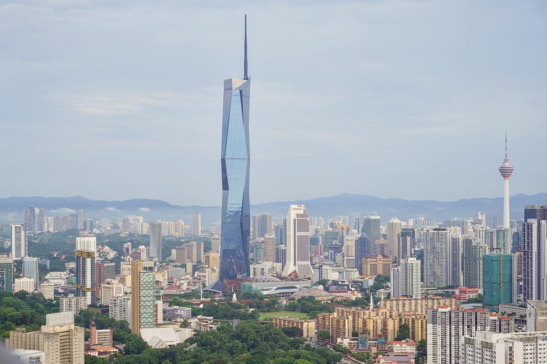 Tòa nhà chọc trời Merdeka 118 vươn lên trên bức tranh toàn cảnh thành phố Kuala Lumpur, được bao quanh bởi nhiều tòa nhà cao tầng, cây xanh và những ngọn núi xa xa vào một ngày có mây rải rác.