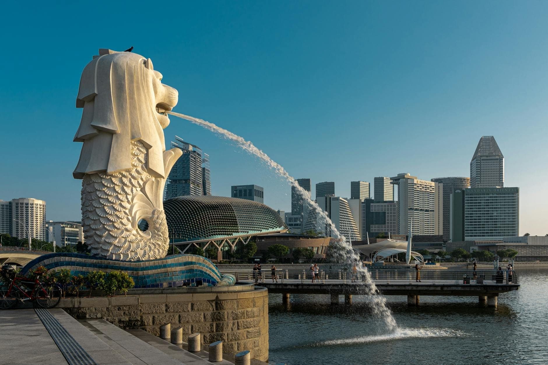 Merlion statue at Merlion Park in Singapore sprays water with city skyscrapers and blue sky in the background, waterfront promenade visible.