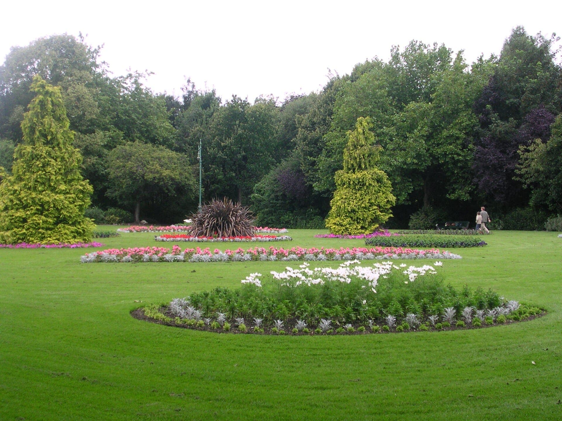 Parterres de flores cuidados y exuberantes praderas verdes en Merrion Square Park, con árboles altos y una persona caminando al fondo.