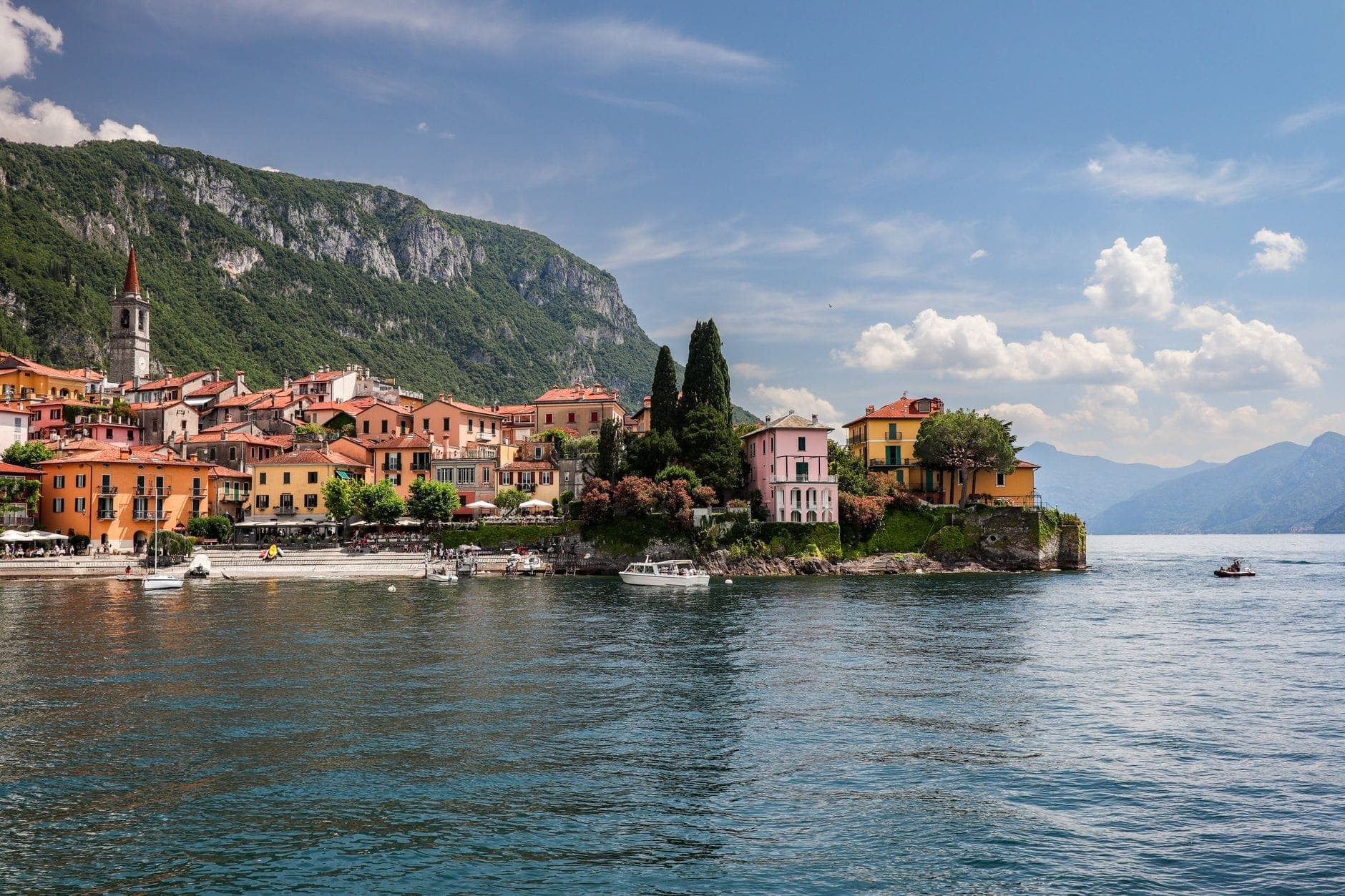 Edificios coloridos y luminosos a orillas del lago en Varenna, en el lago Como, con montañas al fondo y aguas tranquilas en primer plano.
