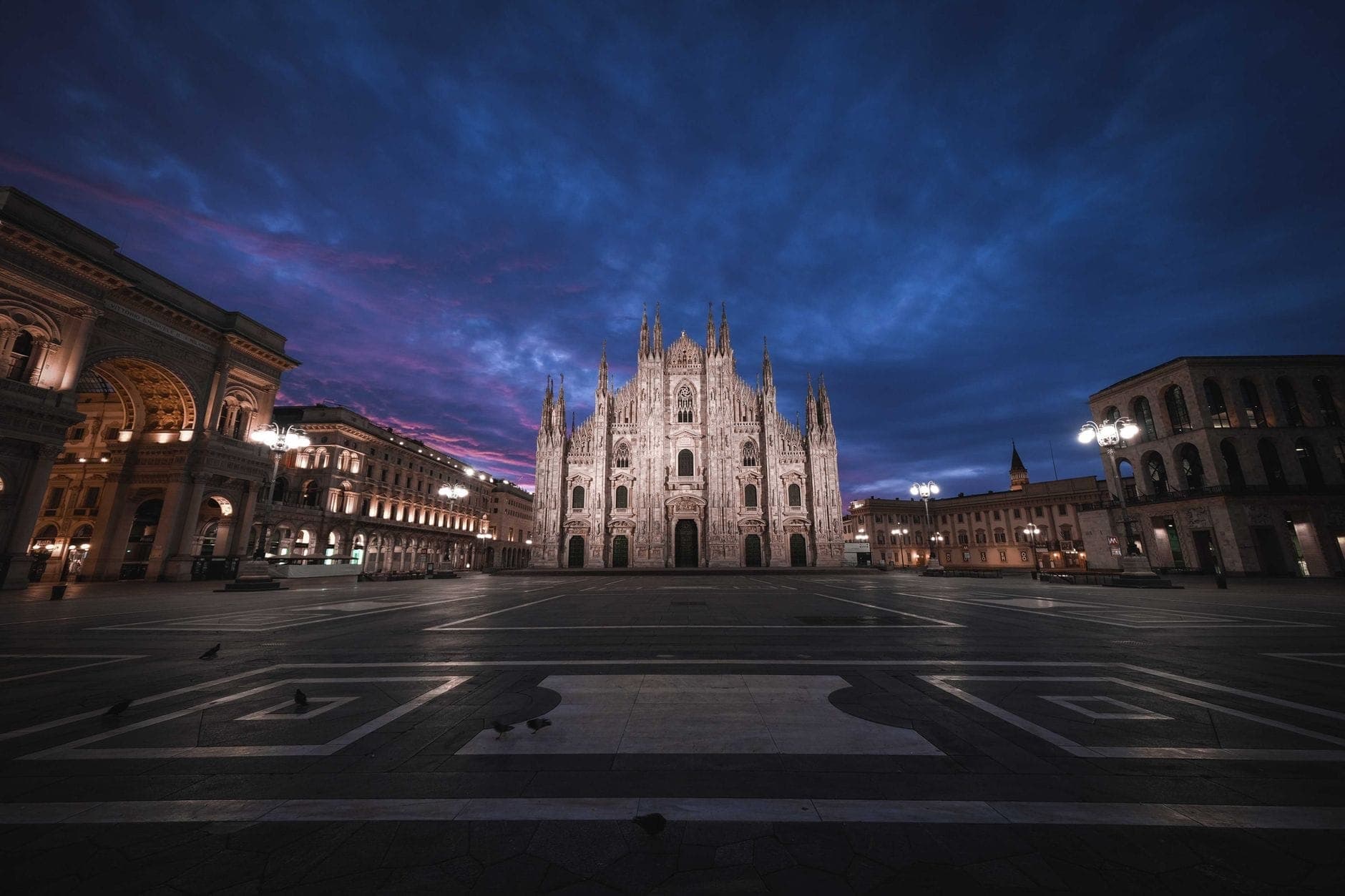 Vista amplia de la Catedral de Milán (Duomo di Milano) y la plaza circundante al amanecer, con un dramático cielo azul y fachada iluminada.