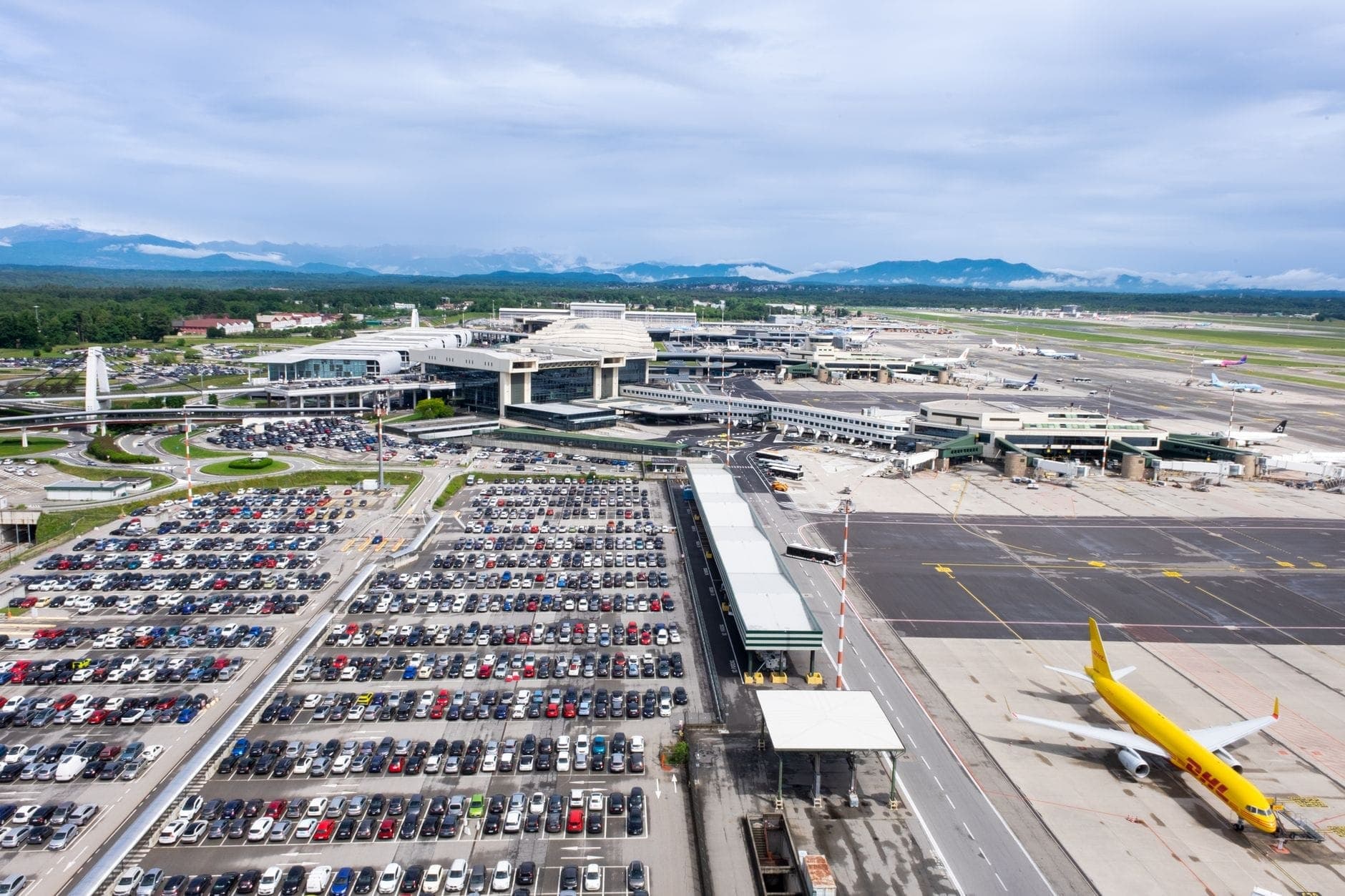 Vista aerea di un grande aeroporto con più terminal, aerei parcheggiati e ampi parcheggi, con uno sfondo di montagne lontane.