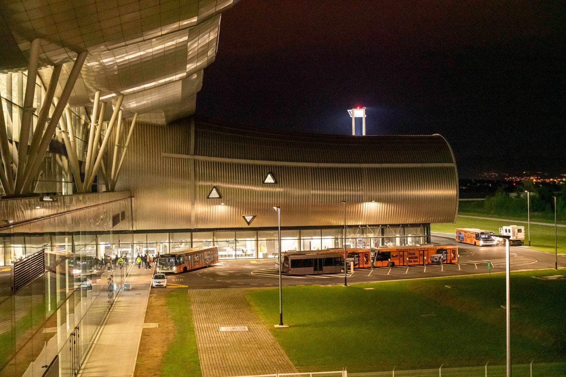 Vista notturna di un moderno terminal aeroportuale a Milano con autobus navetta in attesa all'esterno e passeggeri che si avvicinano all'ingresso.