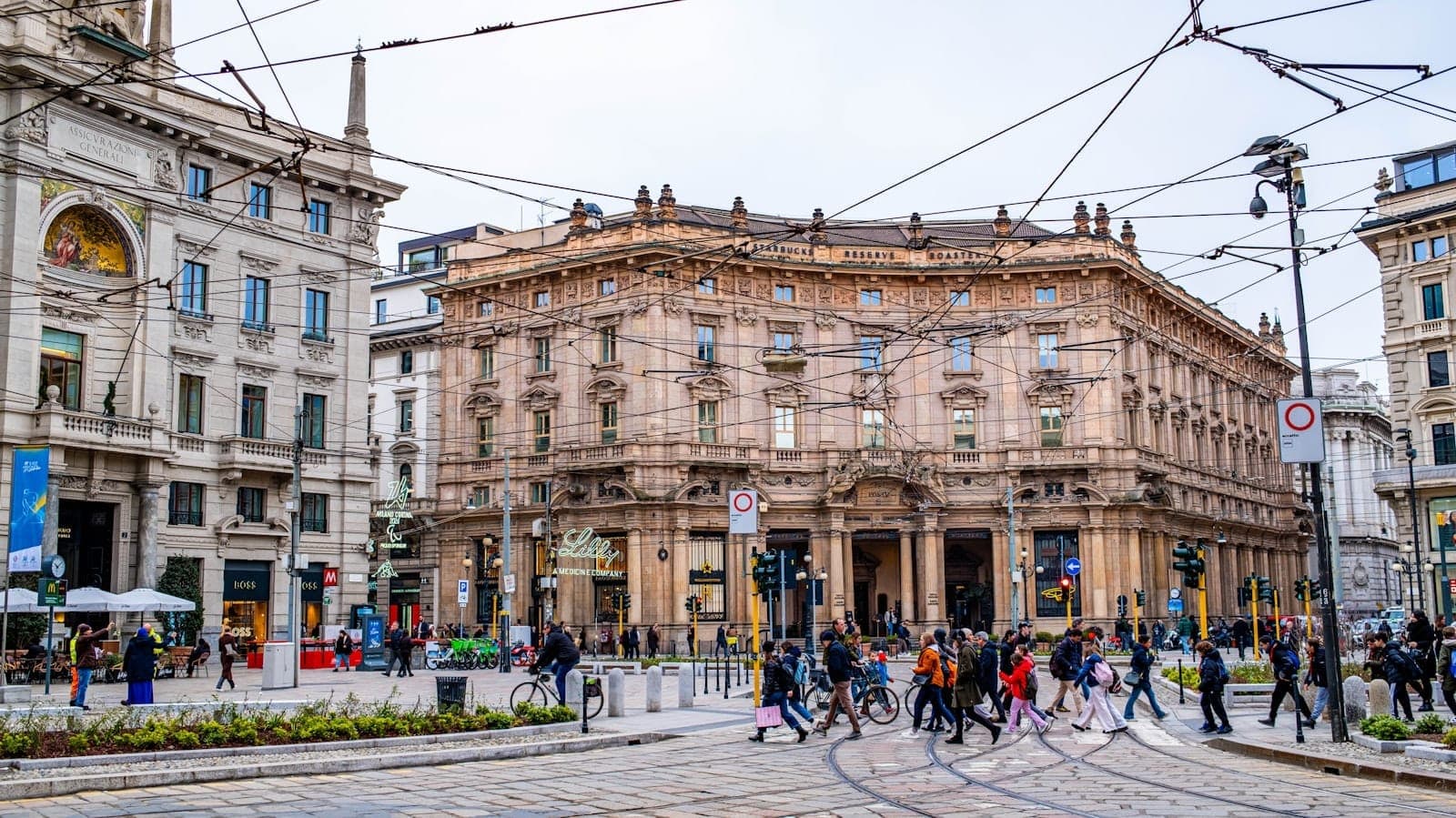 Belebte Kreuzung im Stadtzentrum von Mailand mit historischen Gebäuden, Straßenbahnschienen und Fußgängern, die die Straße überqueren.