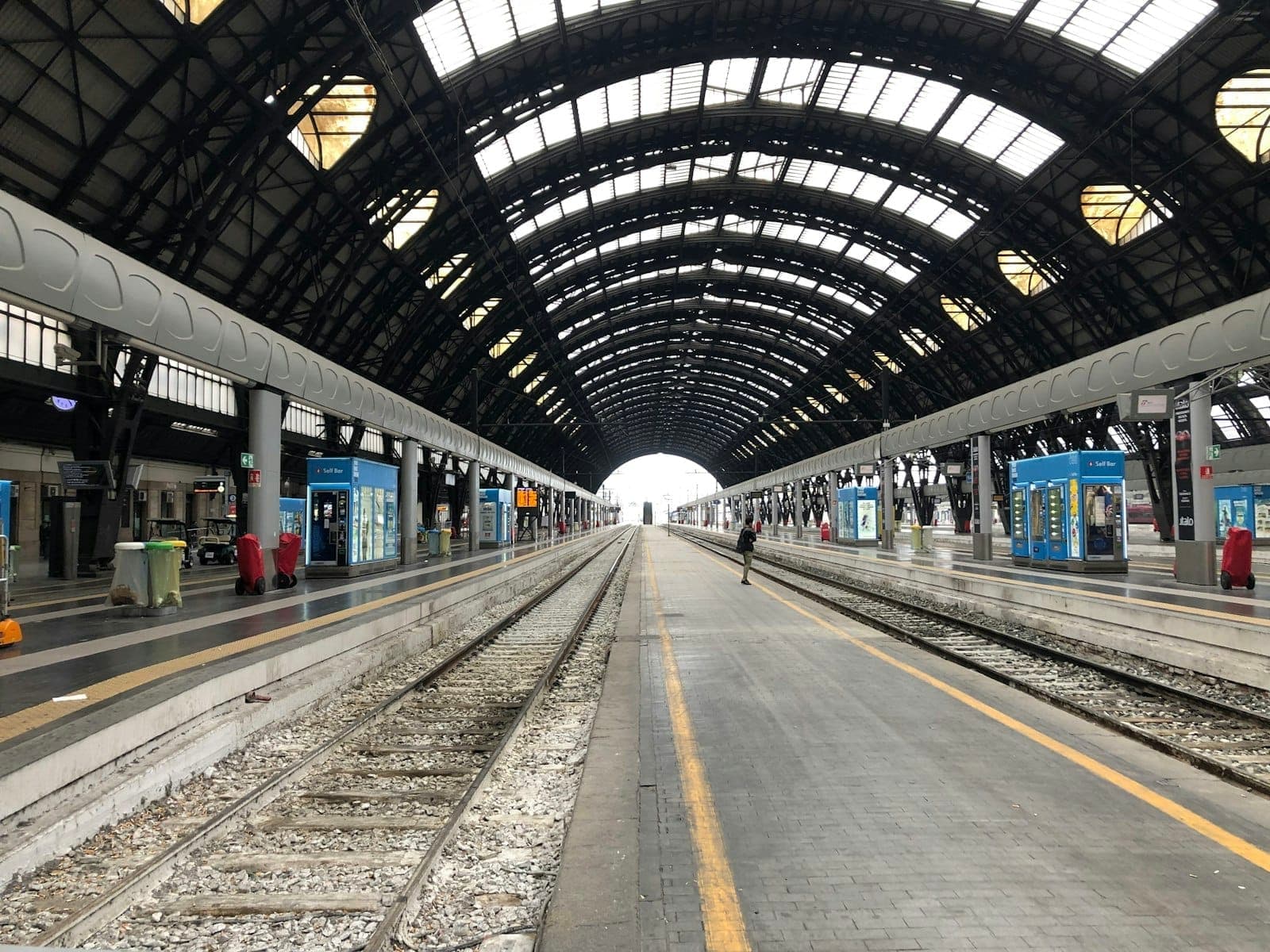 Innenraum des großen Mailänder Bahnhofs mit gewölbtem Stahl-Glas-Dach, leeren Bahnsteigen und modernen Kiosken