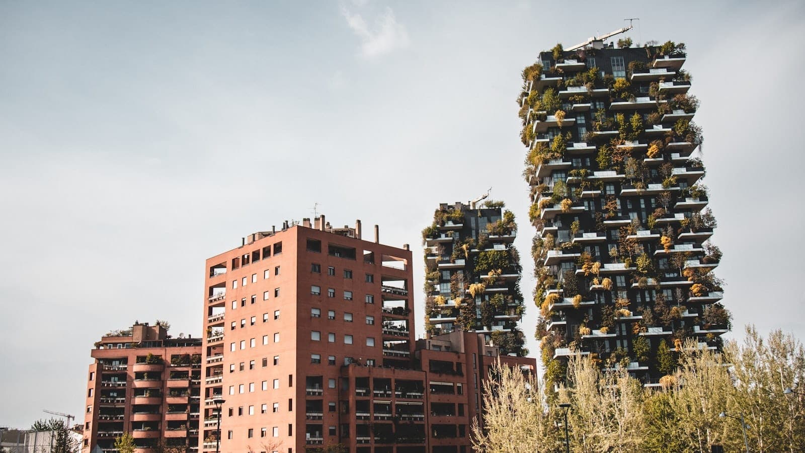 Weitwinkelansicht der Zwillingstürme des Bosco Verticale in Mailand, bedeckt mit Grünpflanzen, neben modernen Backsteingebäuden und Bäumen im Vordergrund an einem klaren Tag.
