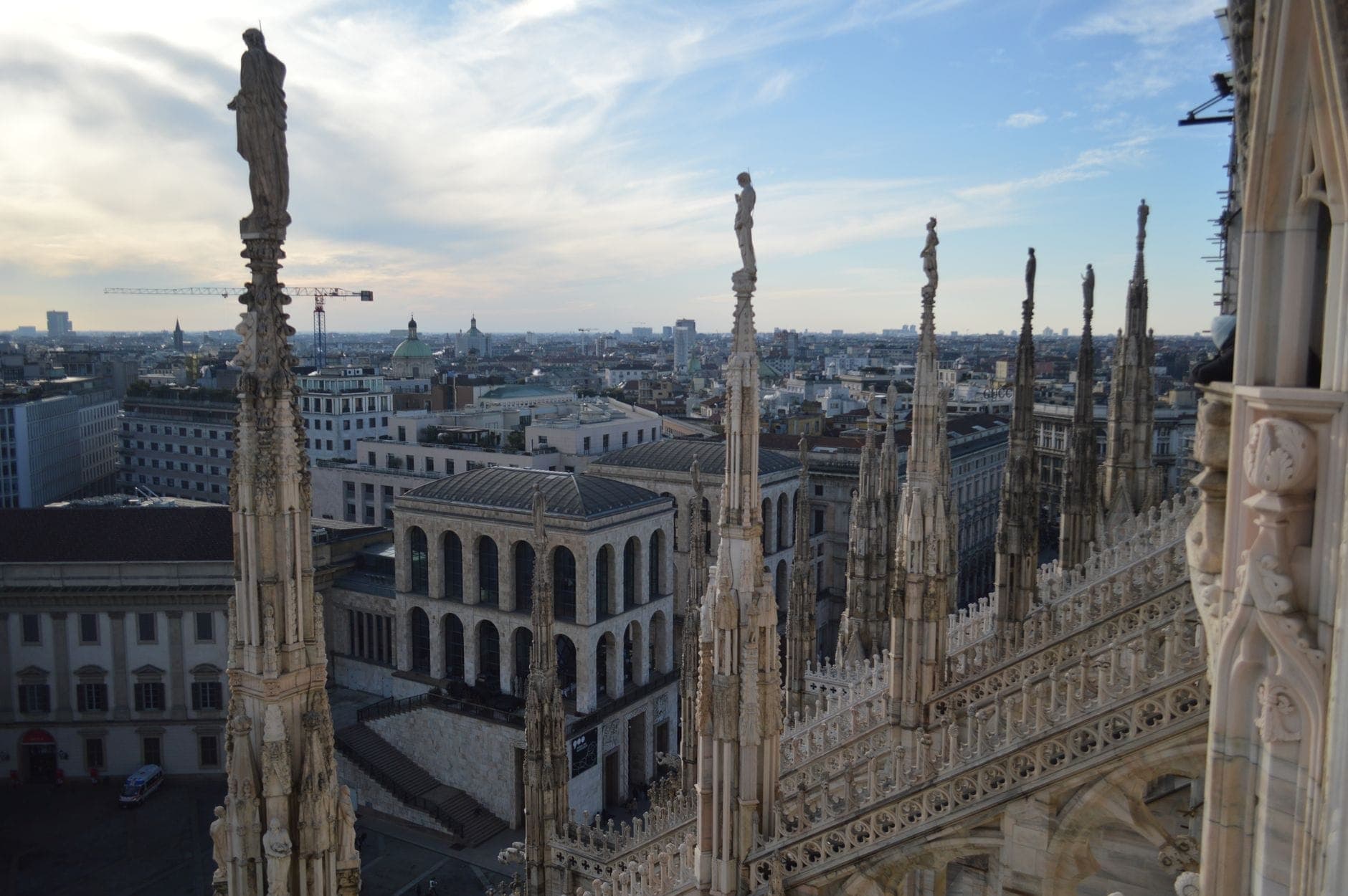 View from Milan Cathedral rooftop terraces showing ornate Gothic spires, marble statues, and the cityscape under a blue afternoon sky.