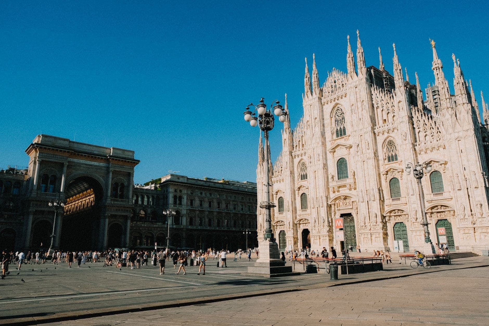 Vista ampia del Duomo di Milano con la Galleria Vittorio Emanuele II e vivaci folle nel quartiere circostante sotto un cielo azzurro limpido.
