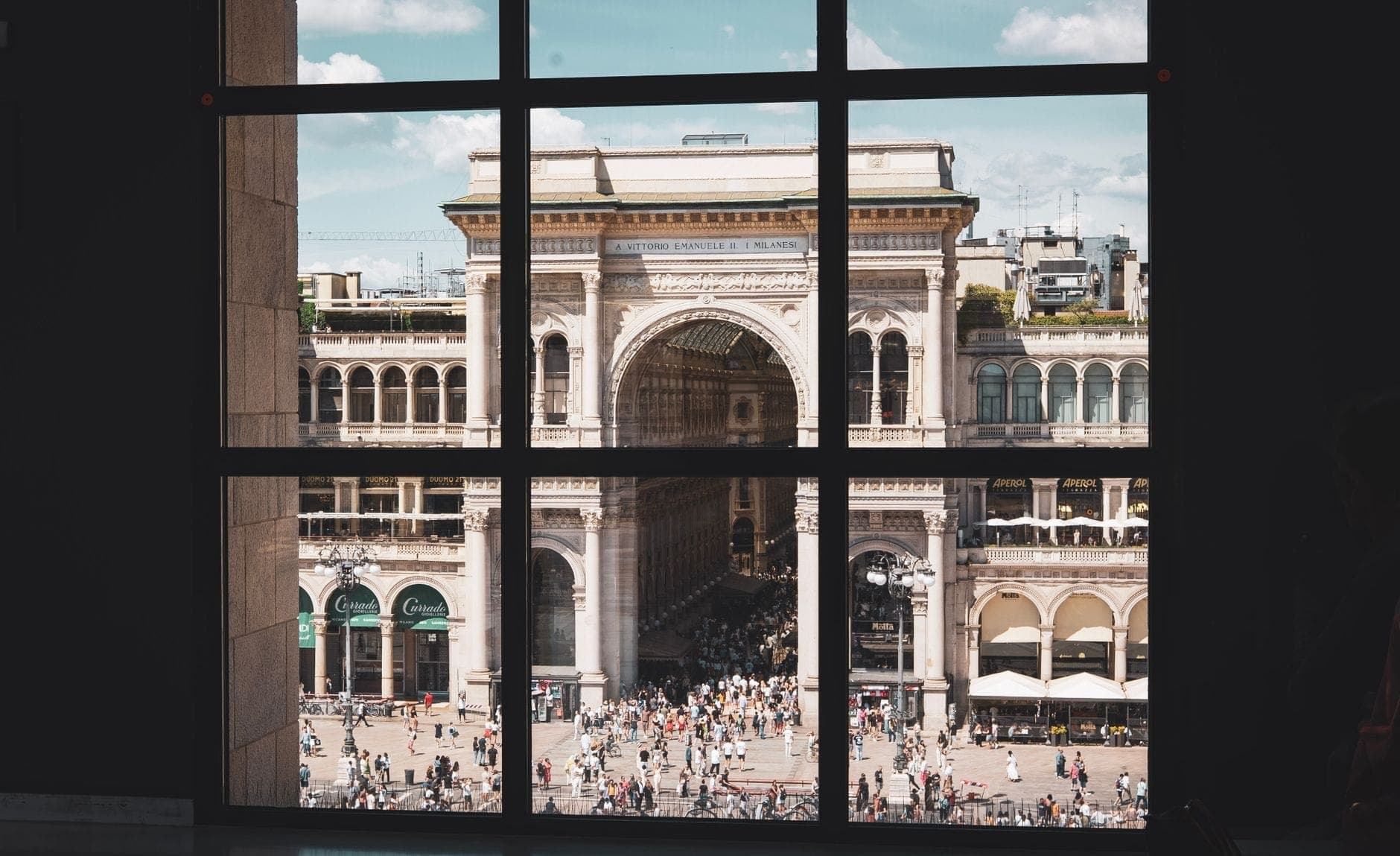Blick durch ein großes Gitterfenster auf die Galleria Vittorio Emanuele II in Mailand, gefüllt mit Menschen und eleganter historischer Architektur.